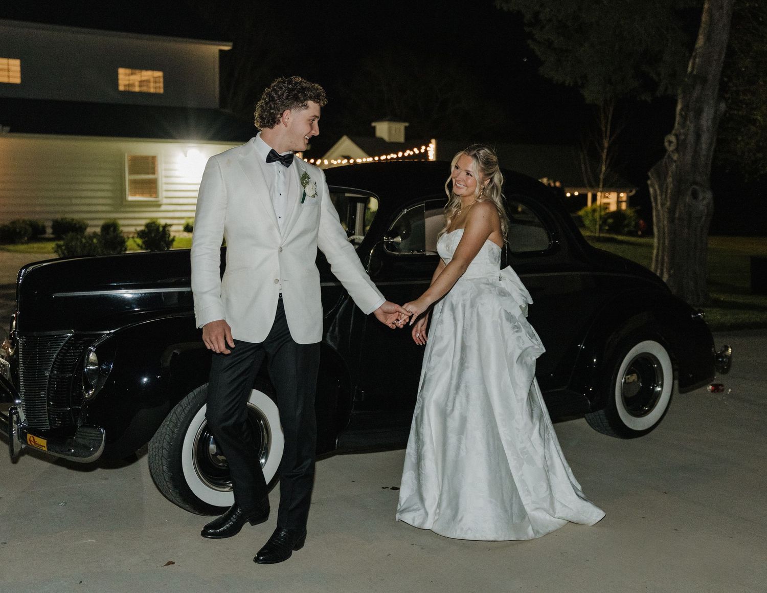 Wedding party poses beside vintage black cars at night with string lights glowing in the background.