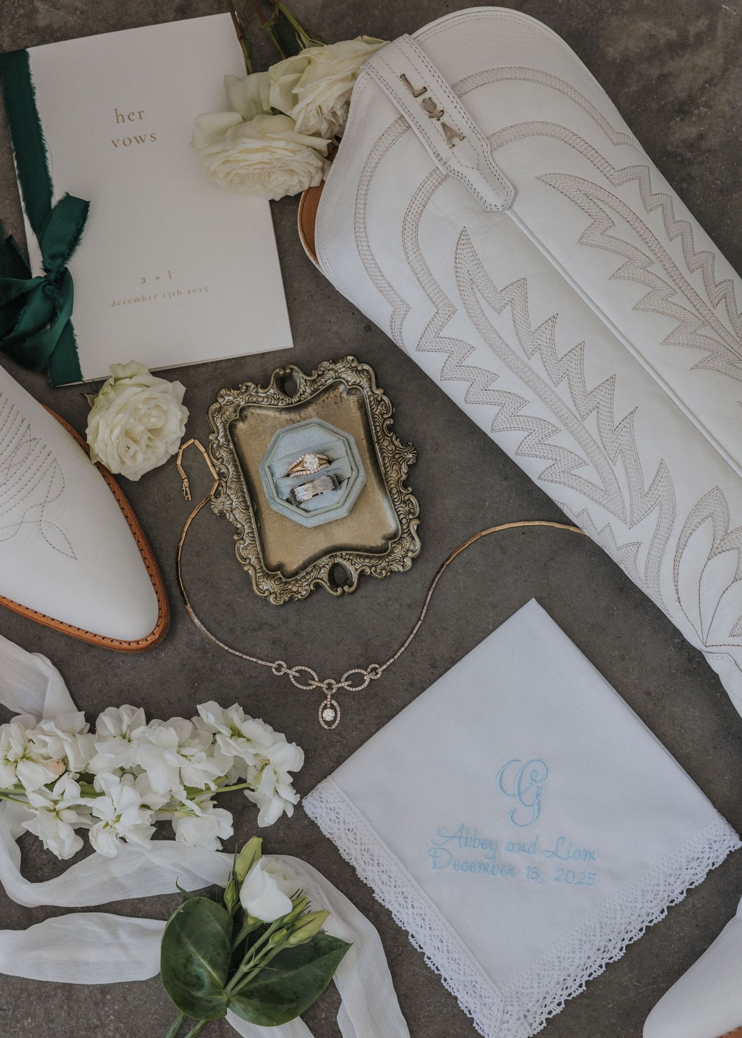 Elegant wedding accessories including embroidered handkerchiefs, vintage frames, and white roses arranged on gray fabric.