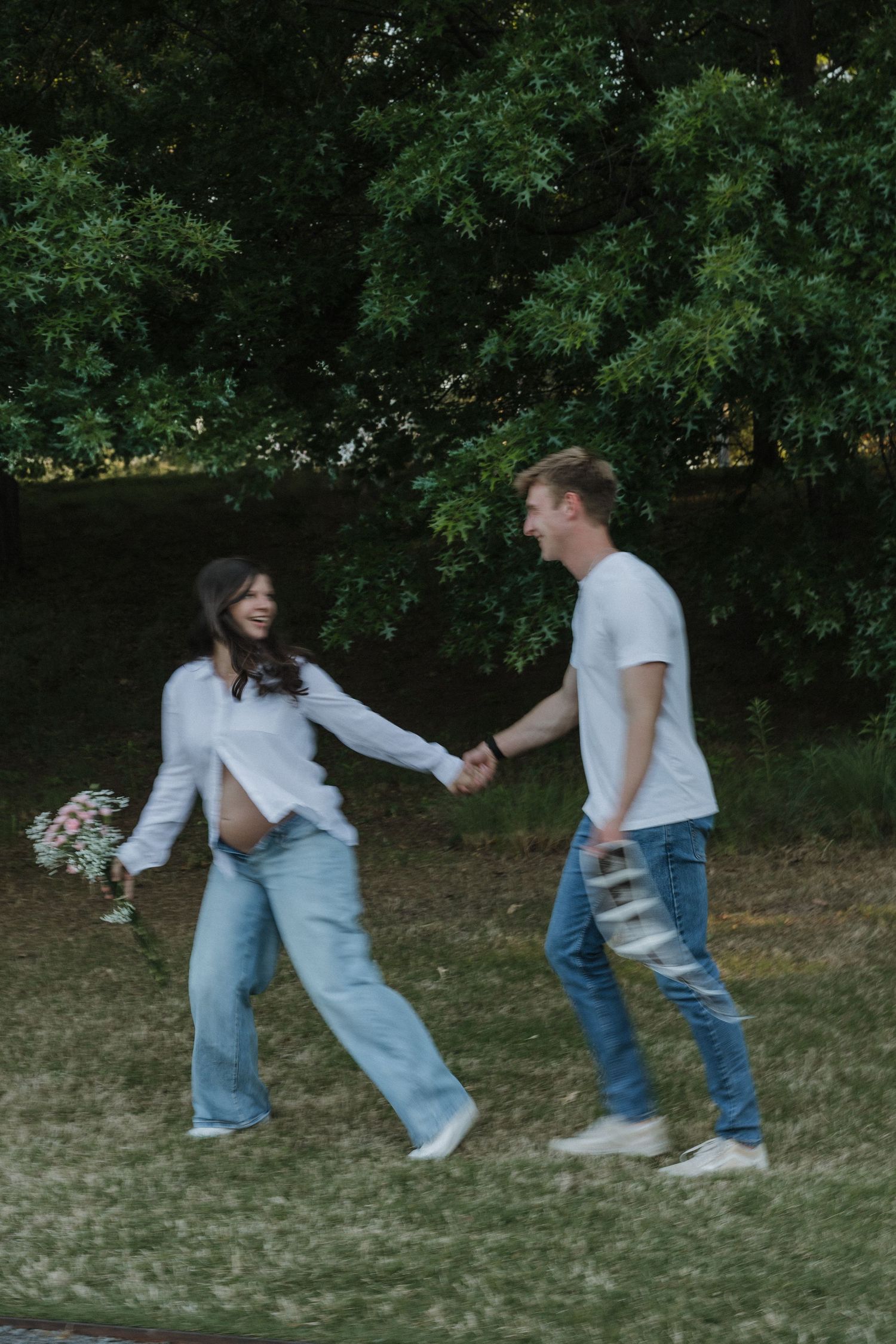 Pregnant woman and man holding hands in a park, both wearing white tops and jeans, with flowers and lush green trees.