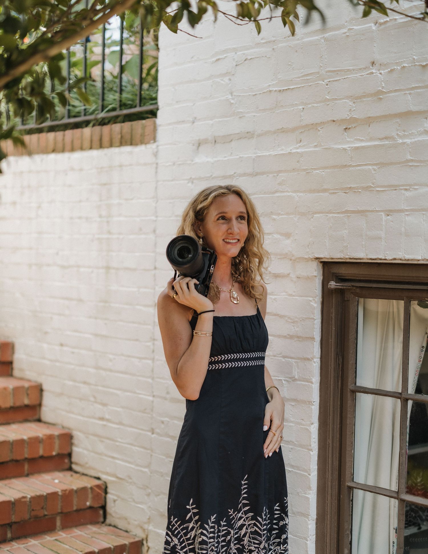 Woman in black floral dress holding a camera outdoors near white brick wall and wooden door.