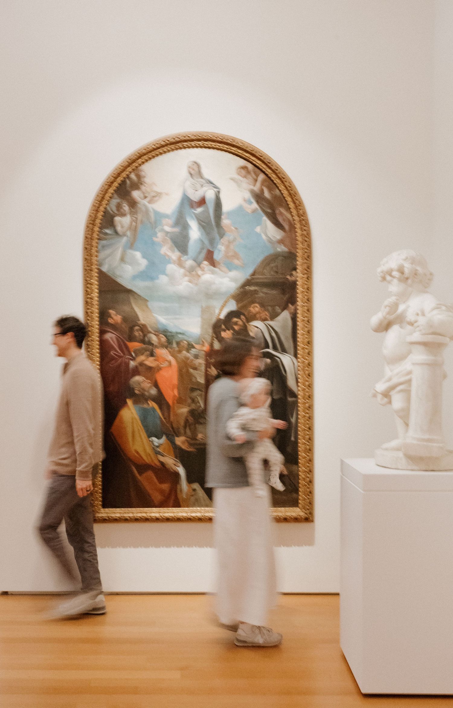Motion-blurred visitors walk past a classical painting in an ornate gold frame displayed on a white gallery wall.