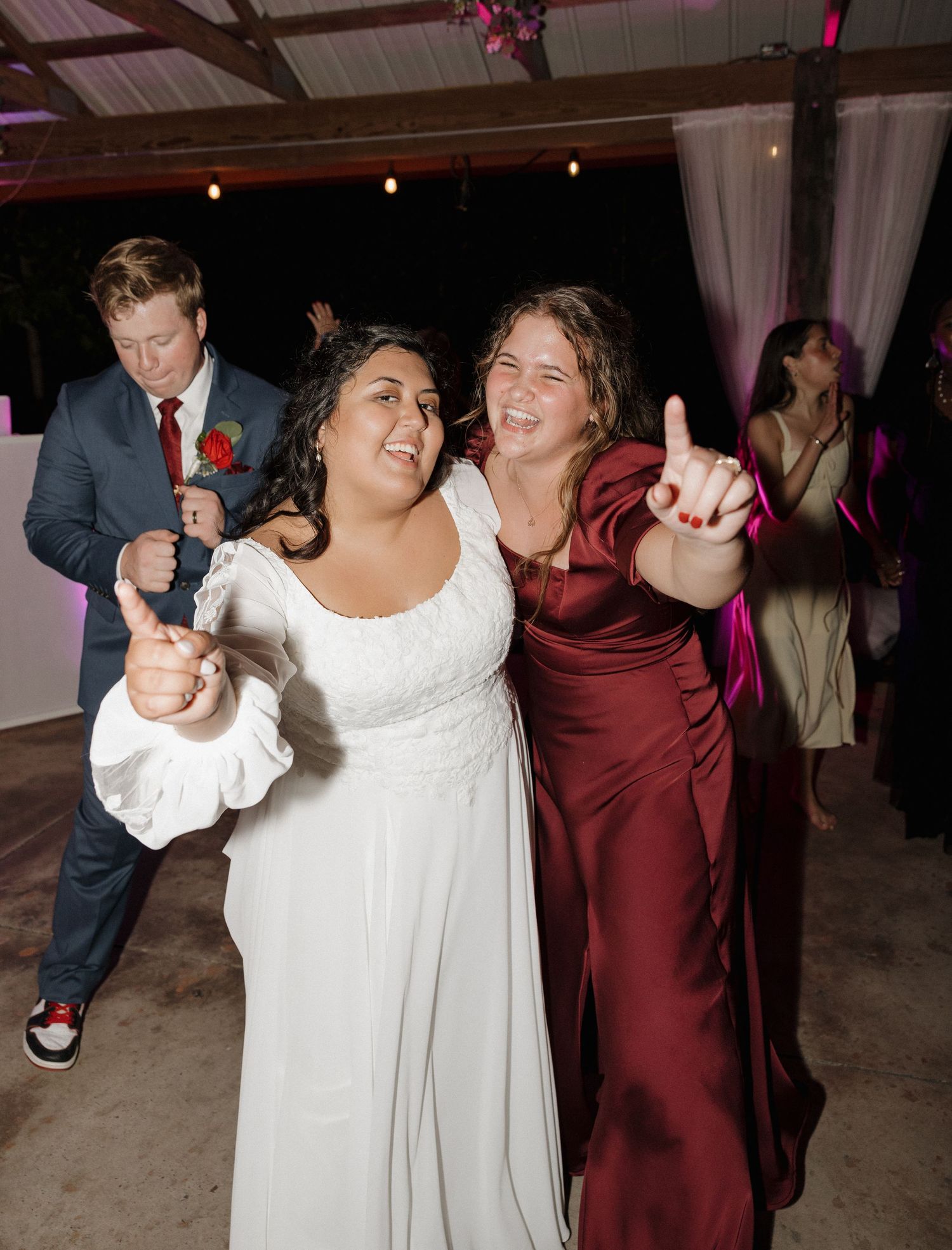 Two wedding guests dancing joyfully together at a reception, one in a white dress and one in burgundy.