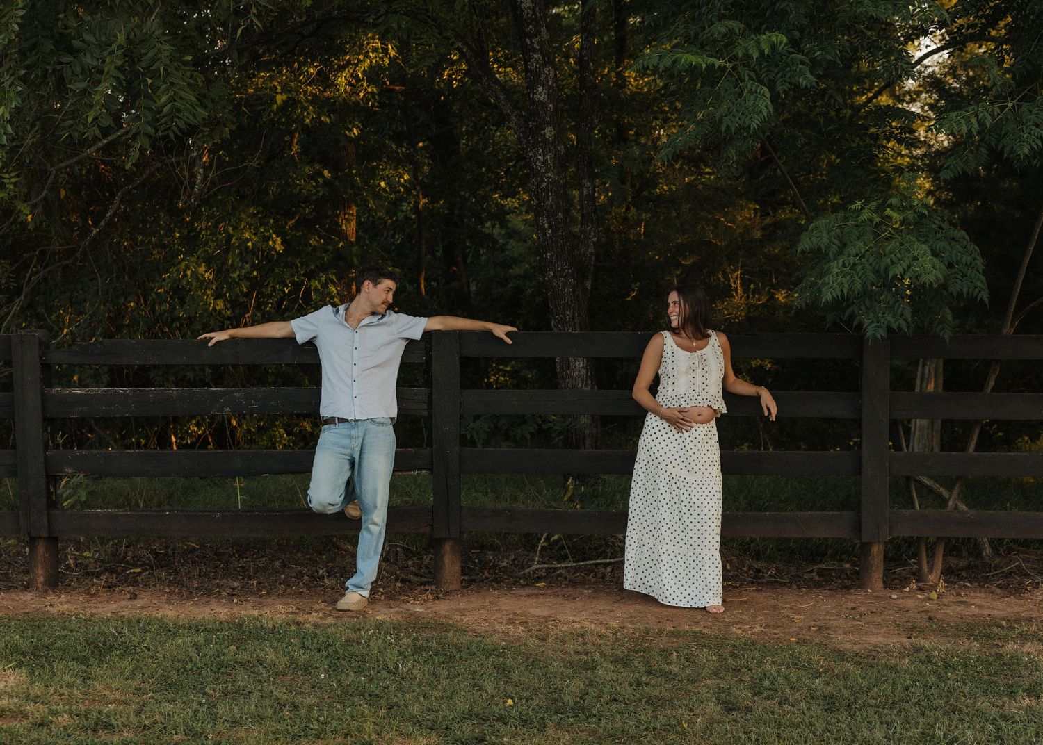 Man balancing on one leg with arms out near wooden fence while woman in white dress stands beside him outdoors.
