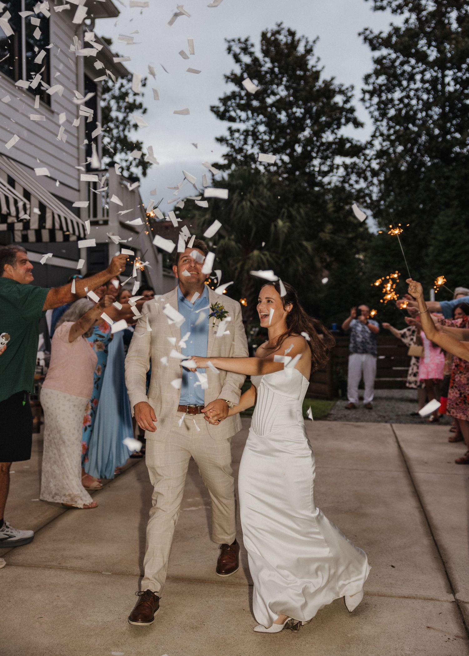 Bride and groom walk through confetti shower with sparklers at outdoor wedding celebration at dusk.