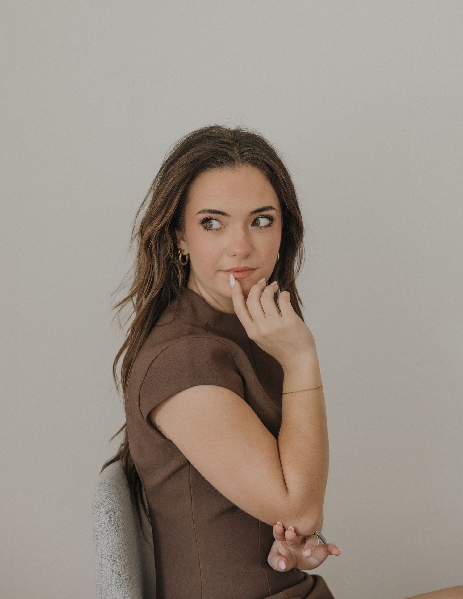 Young woman in brown dress poses thoughtfully with hand near chin, looking over shoulder against neutral background.