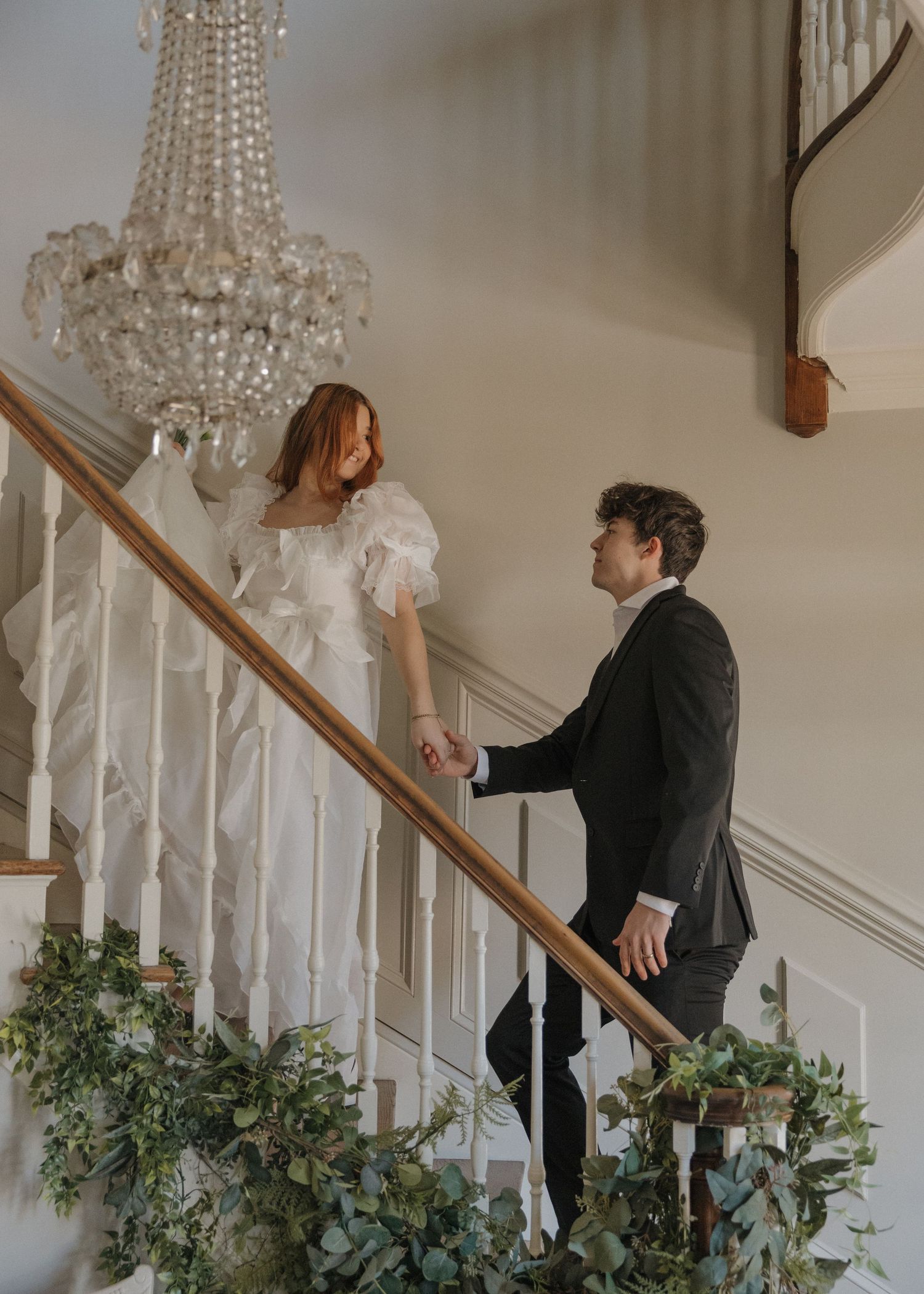 Person in black suit walks up elegant white staircase decorated with greenery and crystal chandelier lighting.