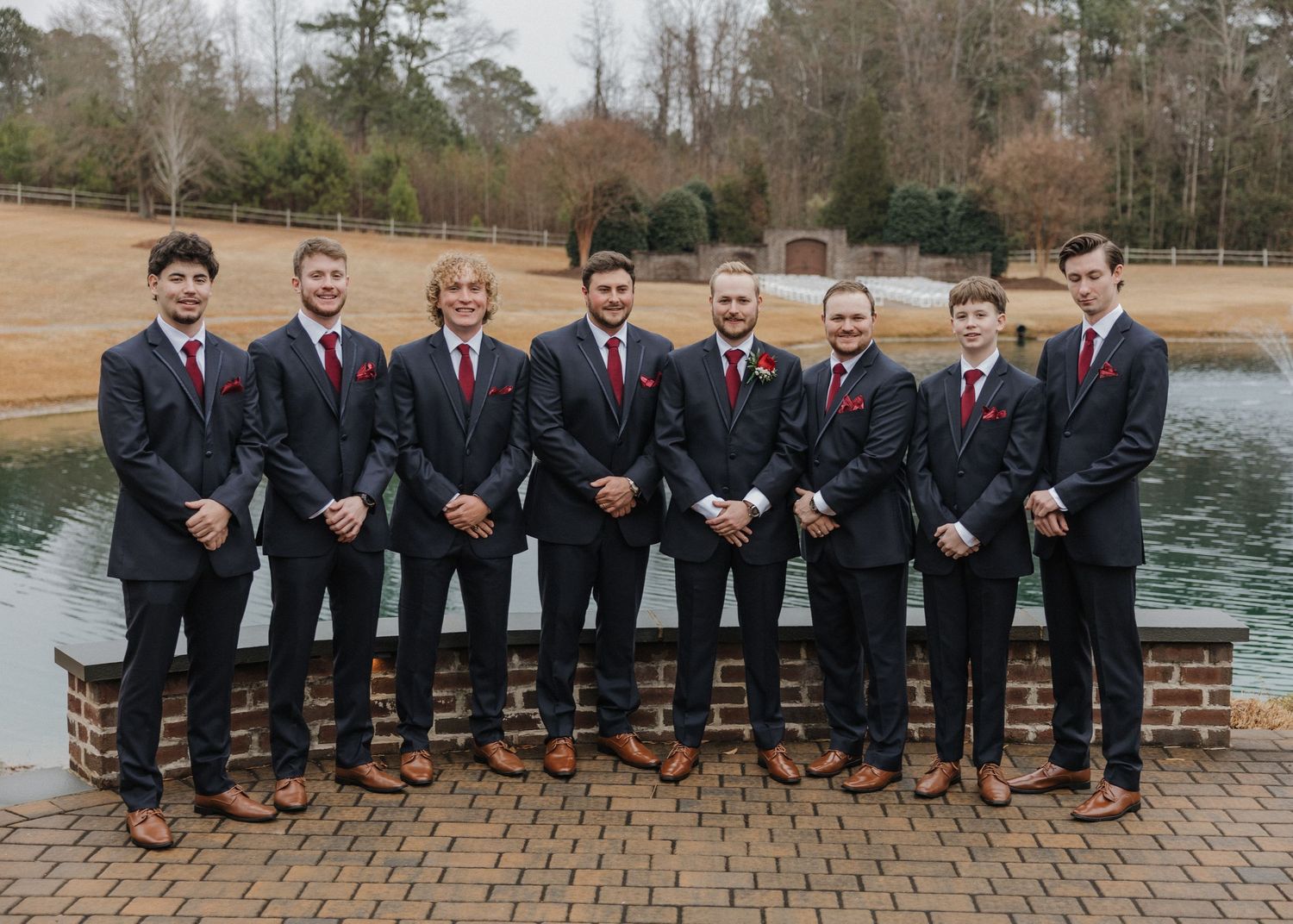 Eight groomsmen in navy suits with red ties pose together outdoors near a pond at a wedding.