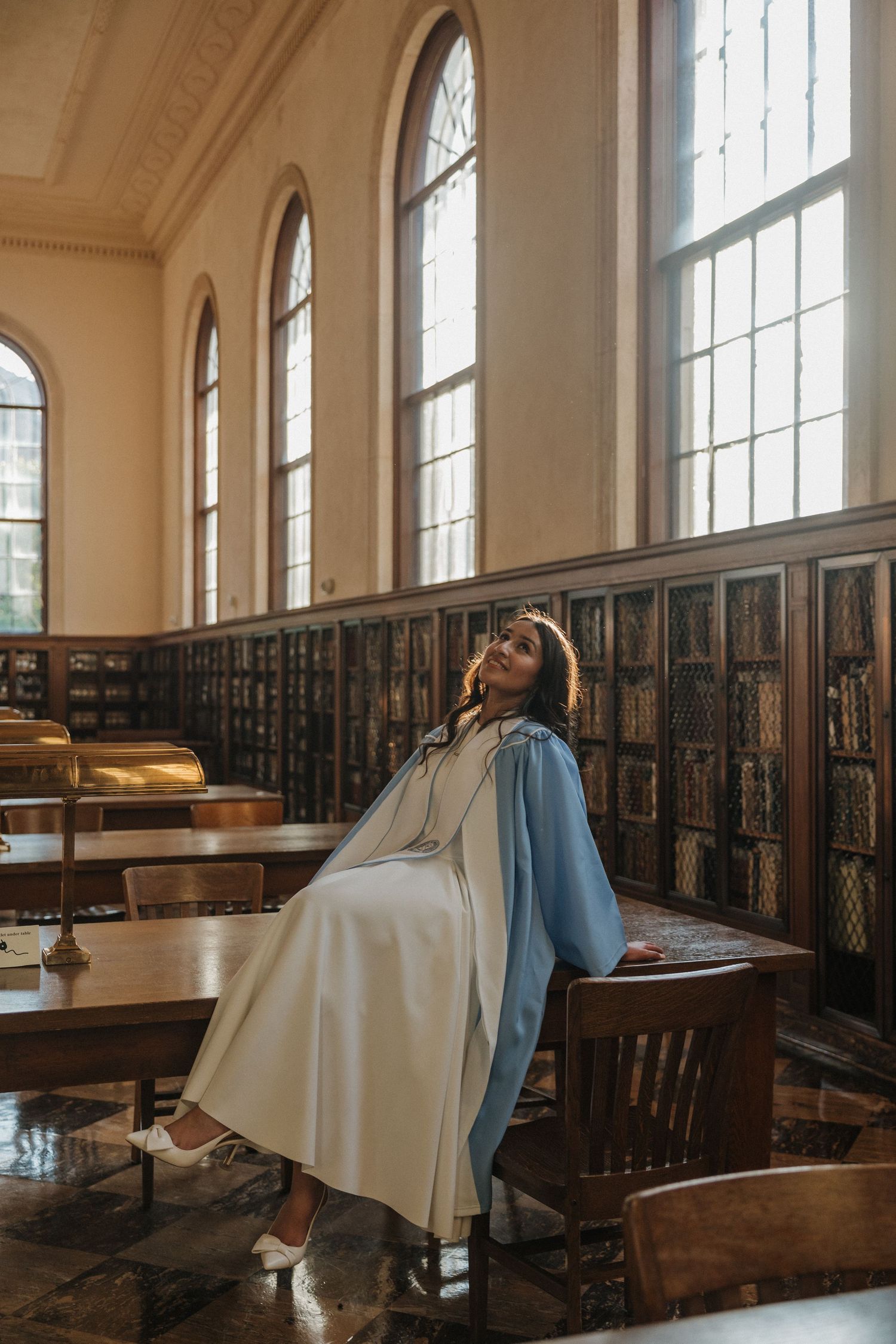 Woman in blue coat and white dress poses elegantly in a grand library with tall windows and wooden bookshelves.