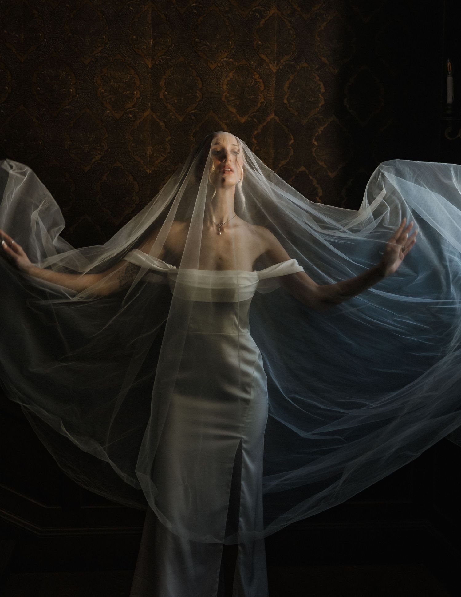 Bride in white strapless gown spreading flowing sheer veil dramatically against dark ornate wallpaper backdrop.