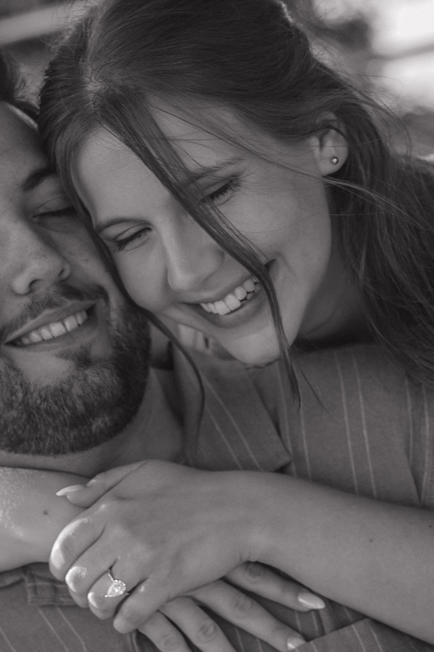 Black and white photo of a smiling couple embracing, woman wearing an engagement ring, sharing a joyful romantic moment.