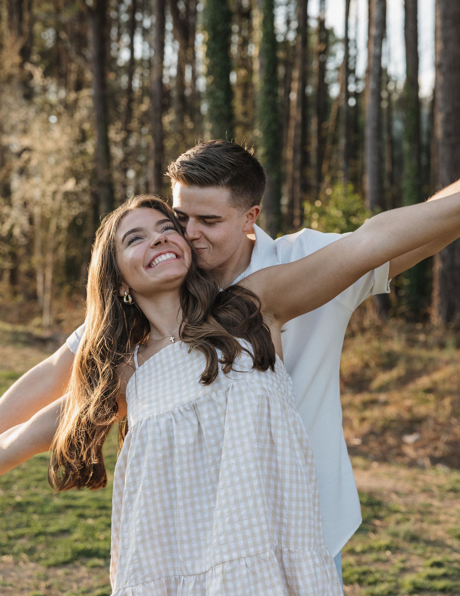 Happy couple embracing outdoors in a forest, woman in white dress smiling with arms outstretched.