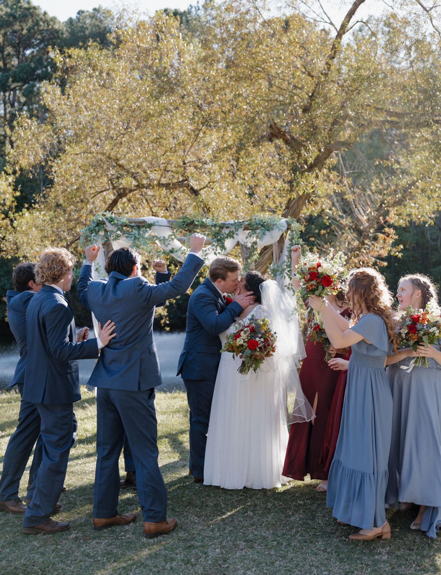 Wedding party in navy and burgundy attire poses under autumn trees.