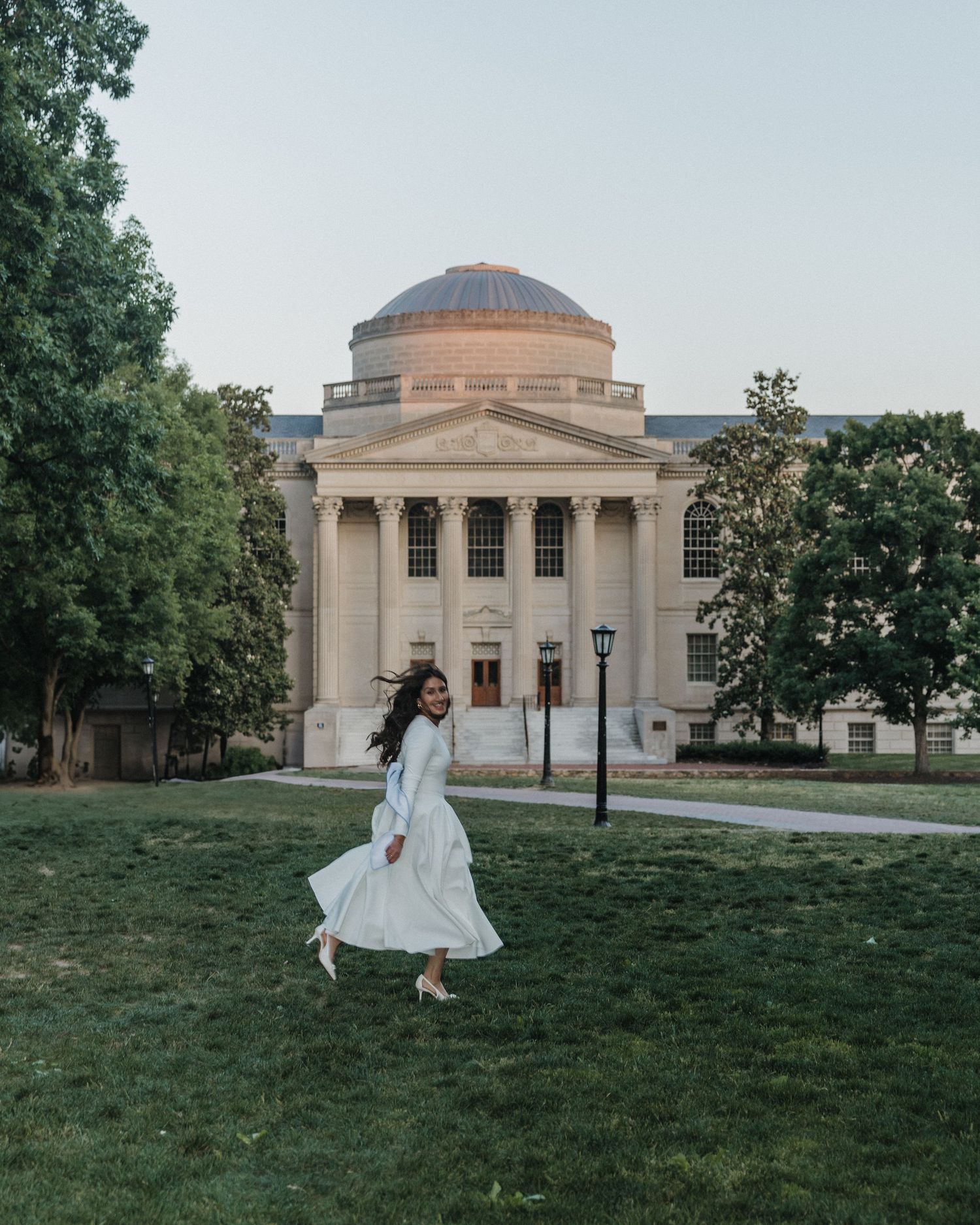 Woman in white dress twirling on green lawn before neoclassical domed building at dusk.