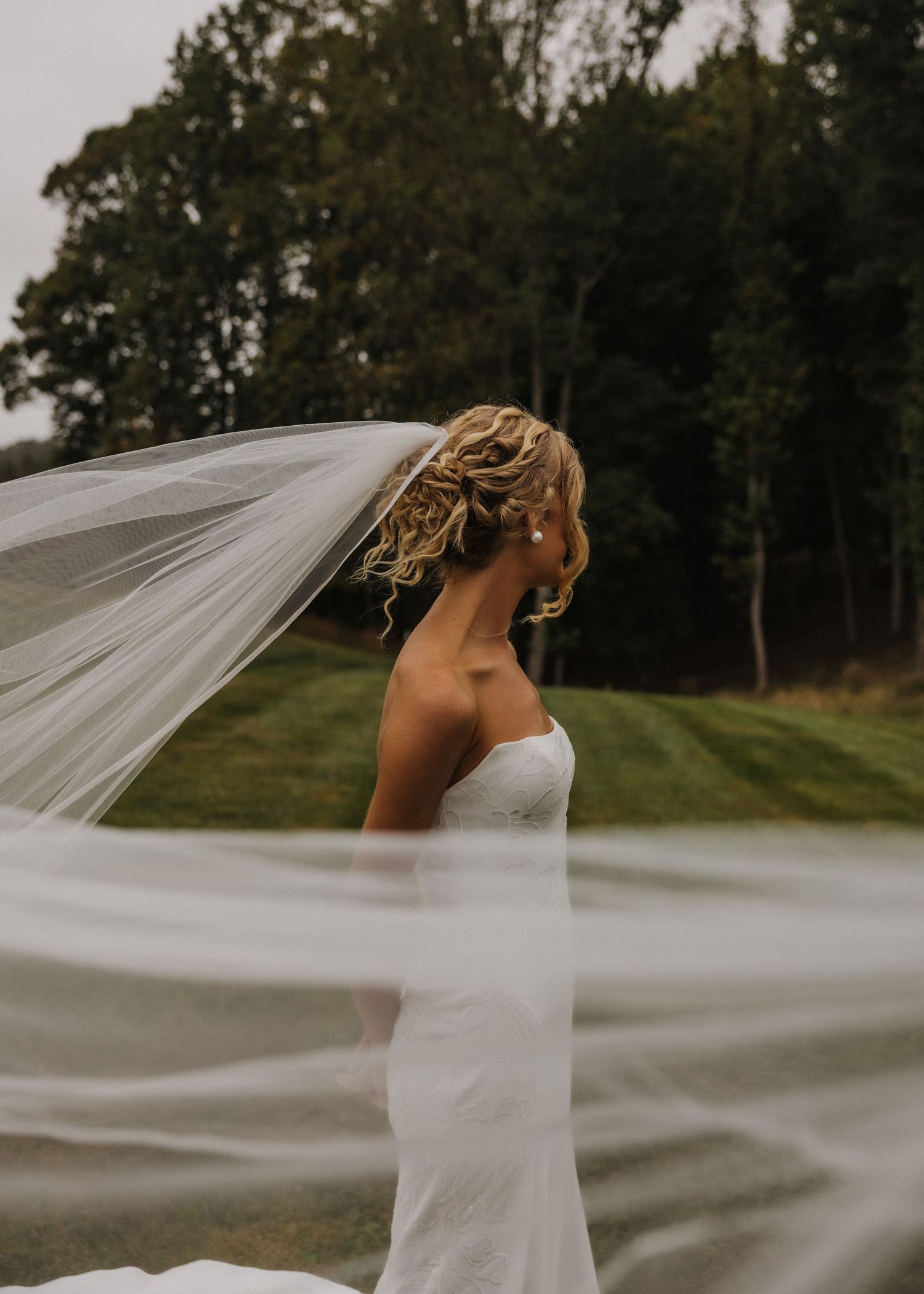 Bride in strapless white gown with flowing veil blowing in wind, standing outdoors with lush green trees background.