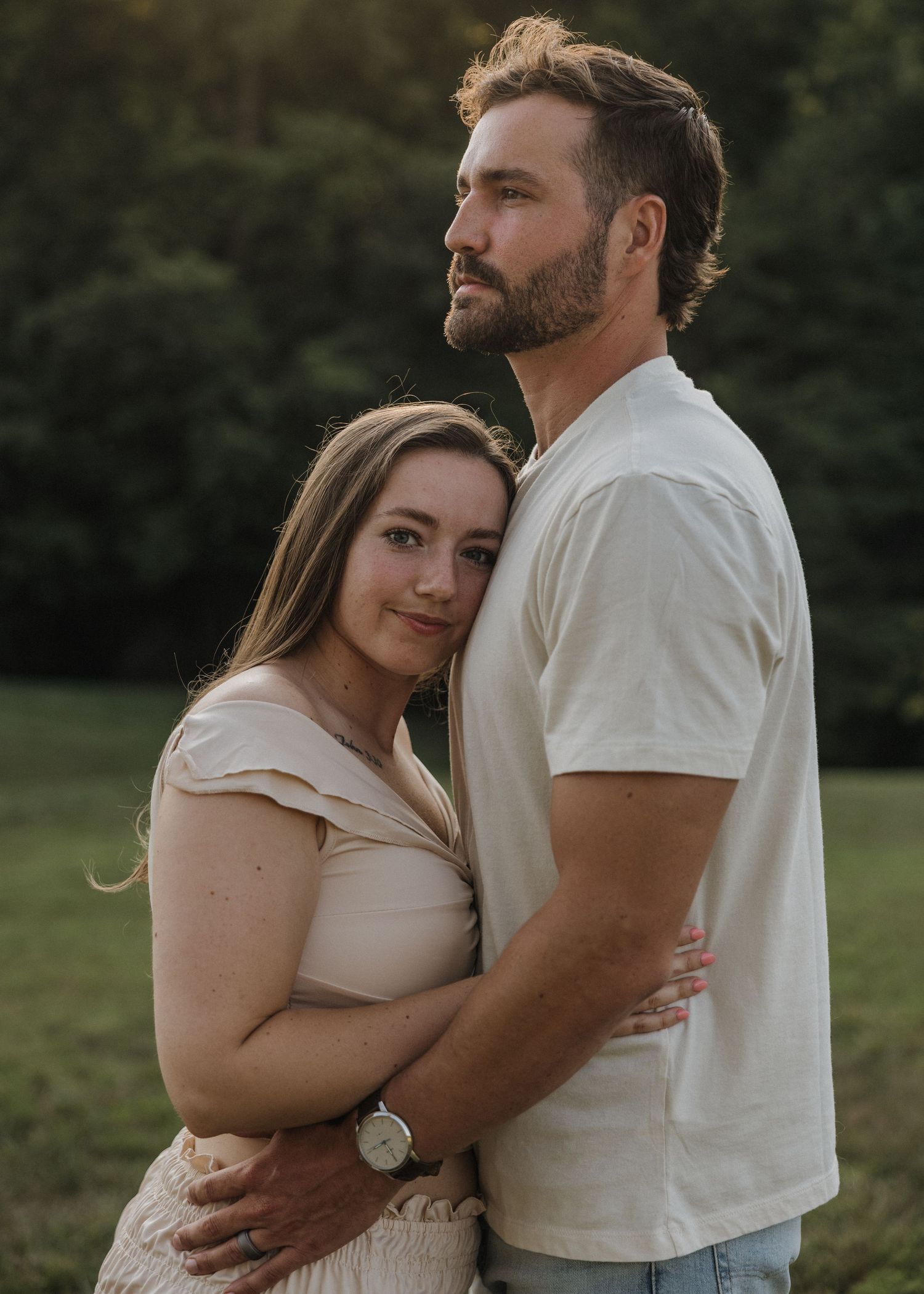 A couple embraces outdoors at golden hour, woman in beige dress leans on man in white tee near lush green trees.