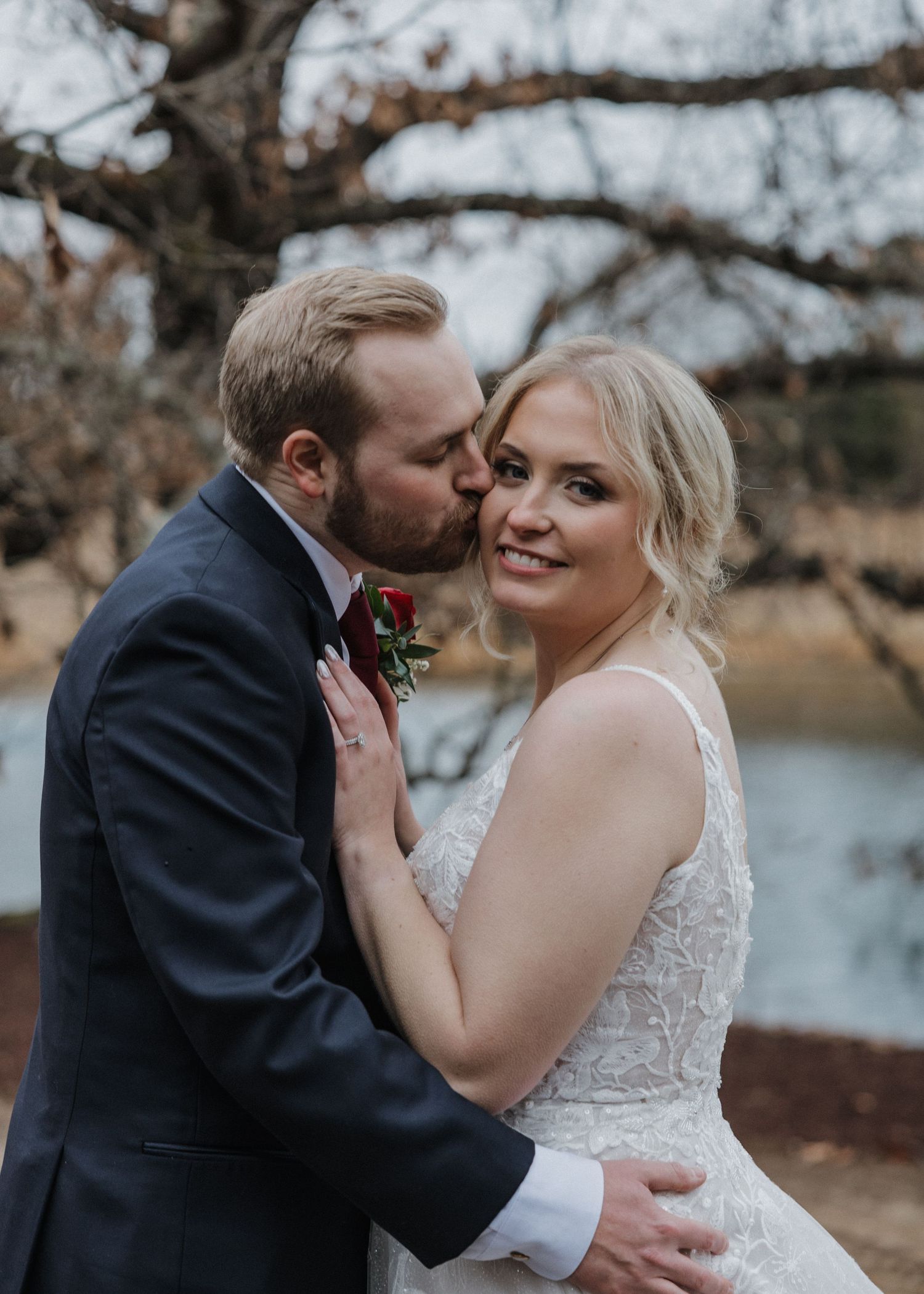 A groom in a navy suit kisses the cheek of his smiling bride in a lace gown outdoors near a pond.