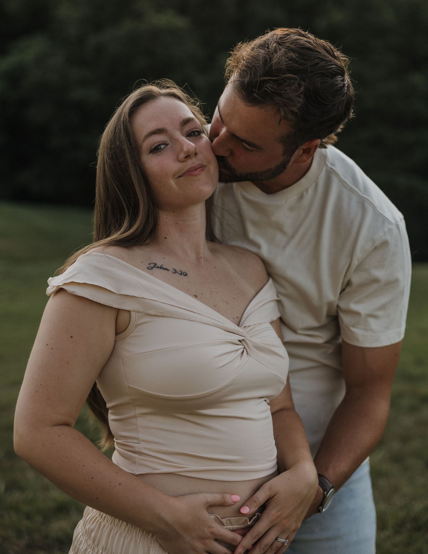 Couple sharing a tender moment outdoors, pregnant woman in beige top with partner kissing her cheek.
