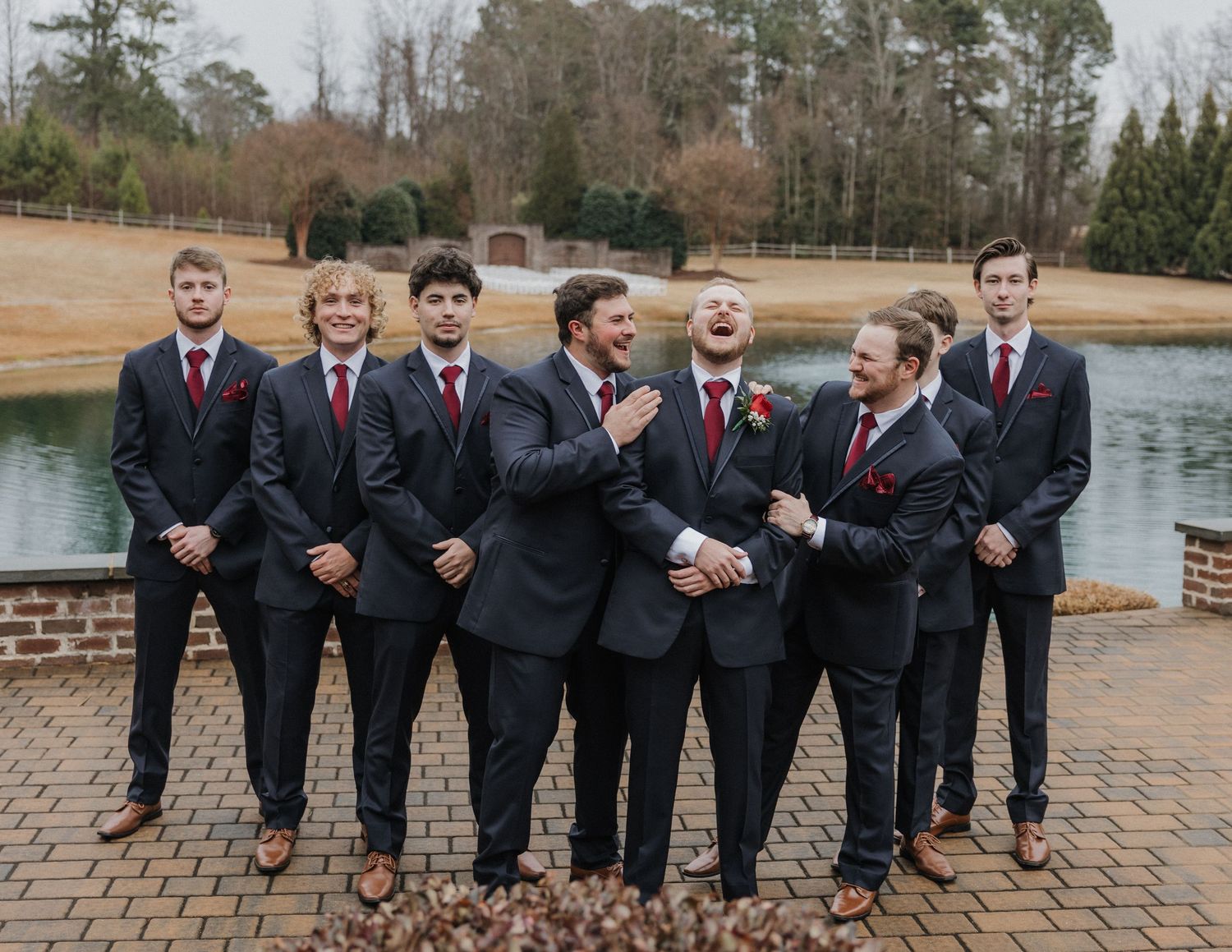 Groomsmen in navy suits and red ties laugh together outdoors near a pond at a wedding.