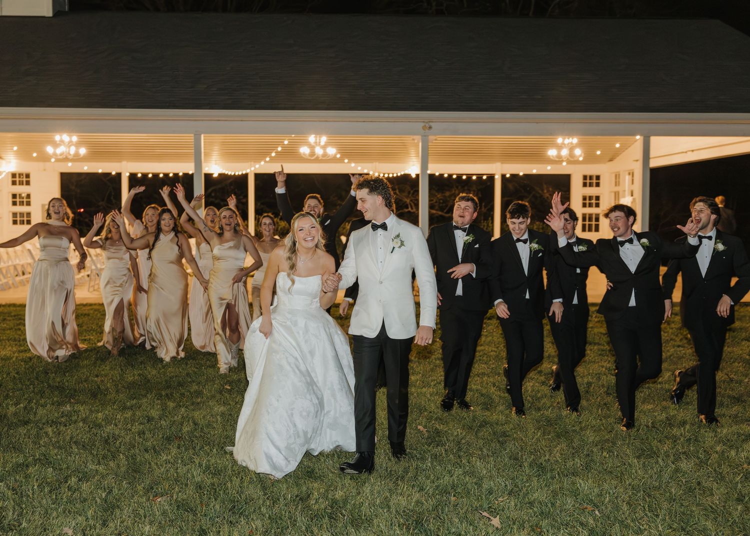 Wedding party celebrating outdoors at night, bride and groom leading bridesmaids in champagne gowns and groomsmen in tuxedos.