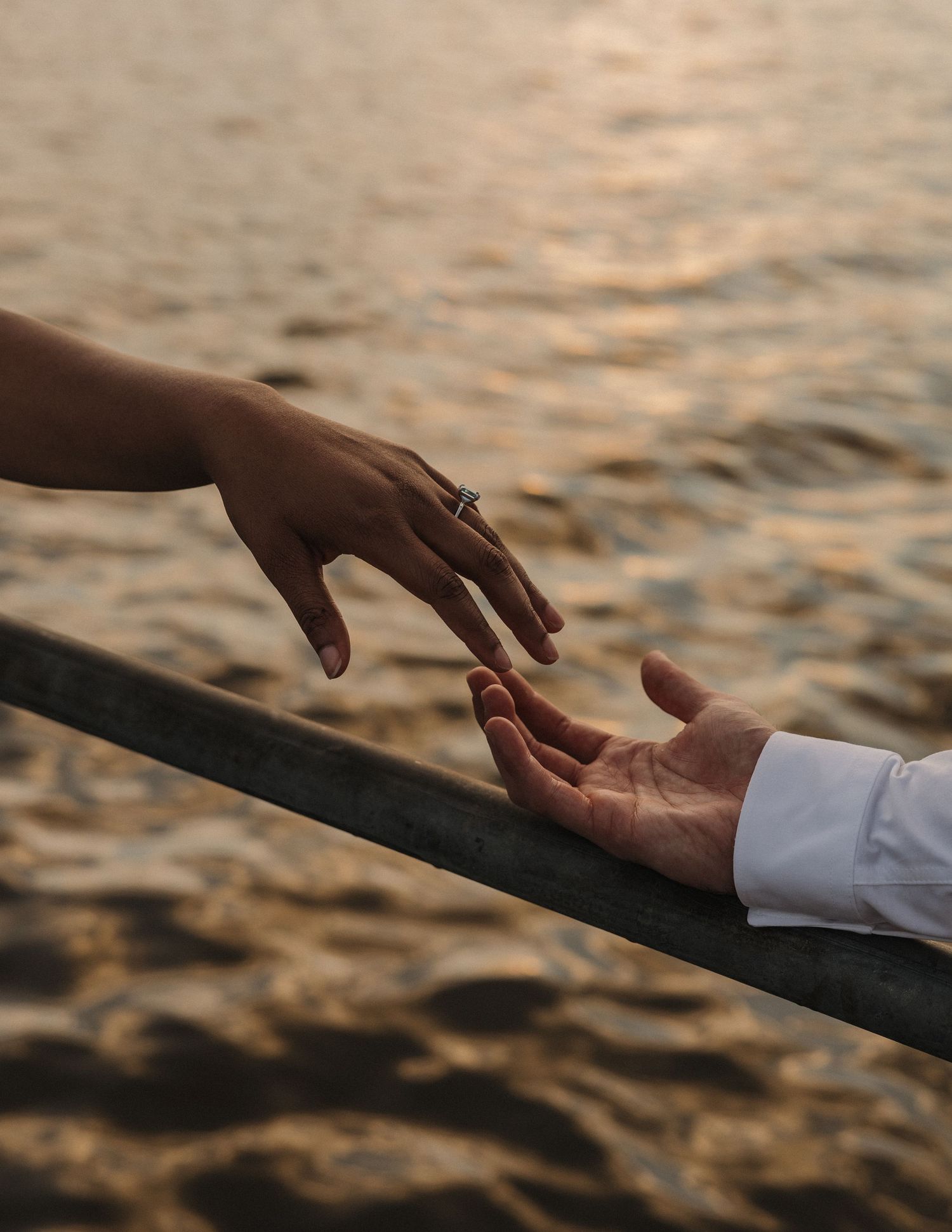 Two hands reaching toward each other over a railing at sunset, one adorned with a diamond engagement ring.