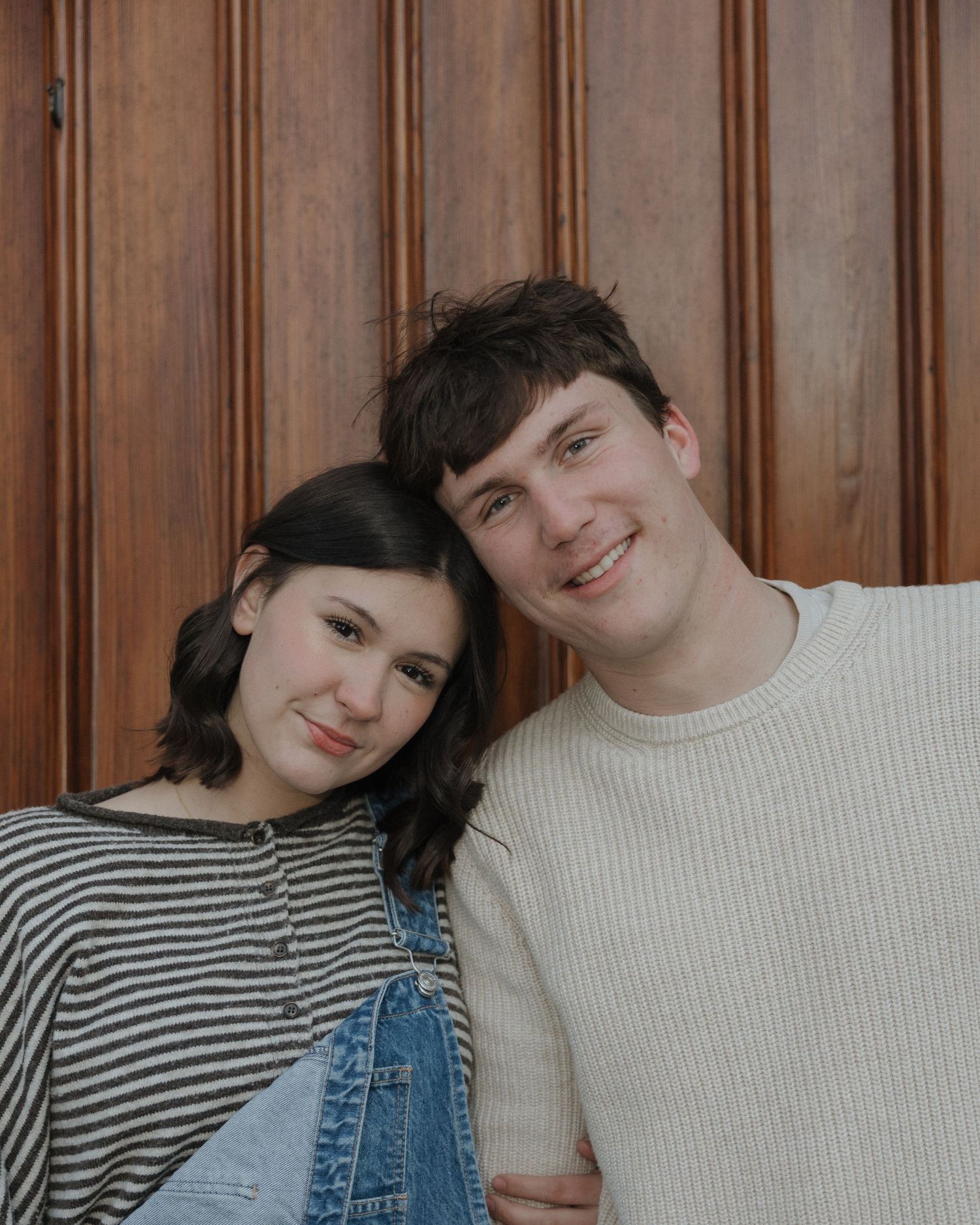 A couple in casual attire poses together against a wooden paneled wall, wearing striped and solid sweaters.