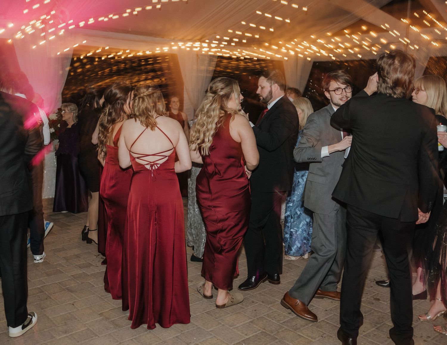 Guests in formal attire dancing at a wedding reception under warm string lights.