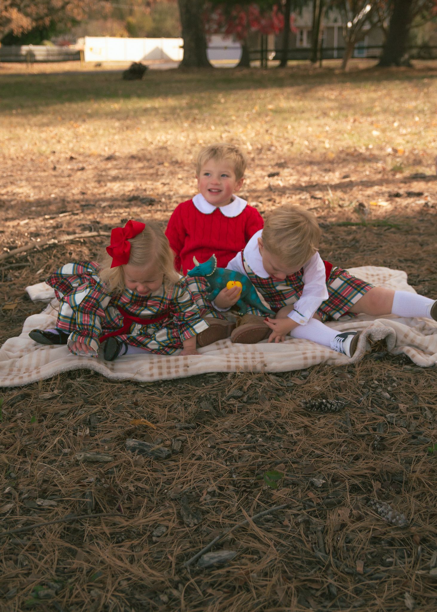 Two children in holiday outfits sit on a blanket with teddy bears wearing matching plaid outfits during an outdoor photoshoot.