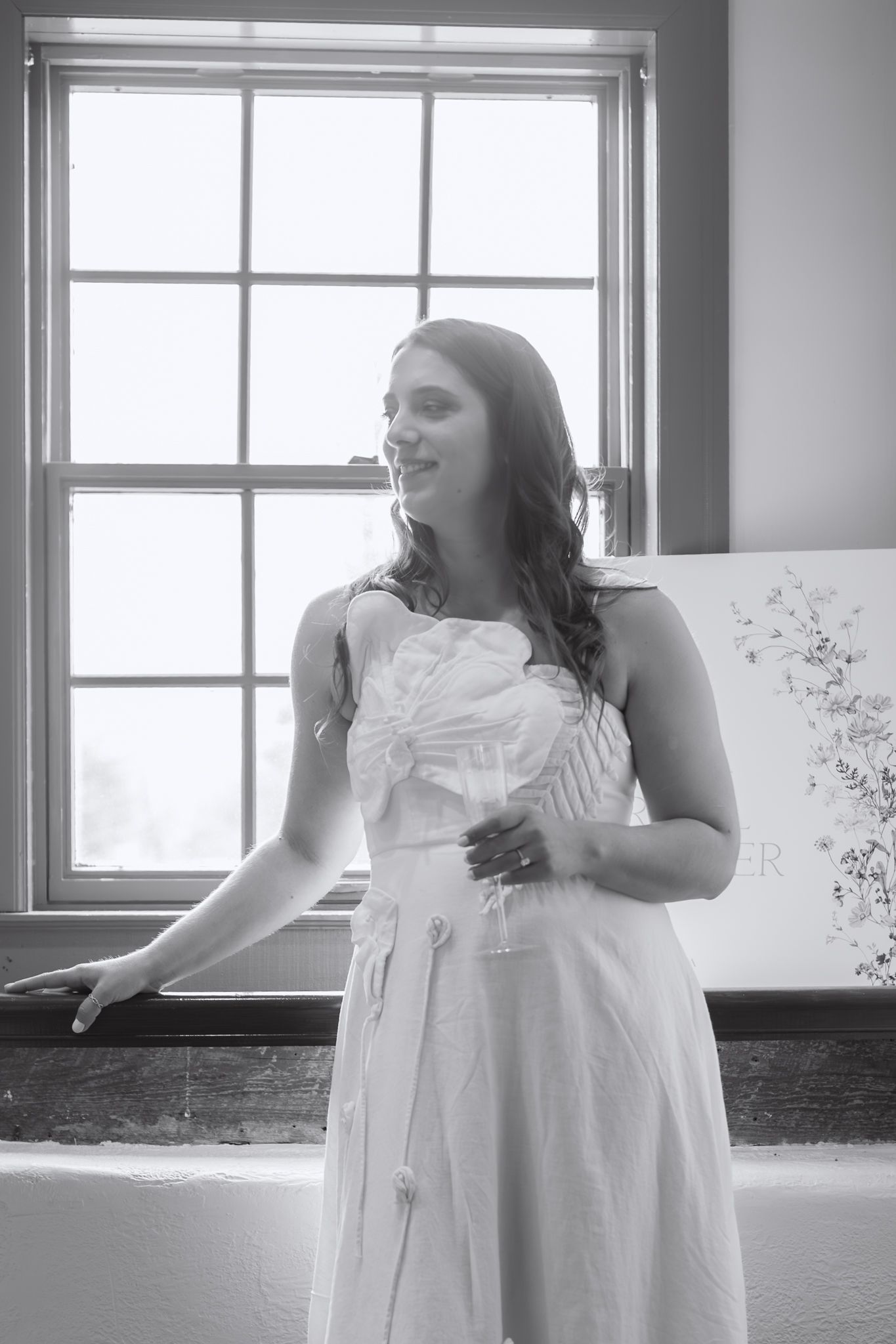 Black and white photo of someone in a flowing white dress standing by a bright window in an interior setting.