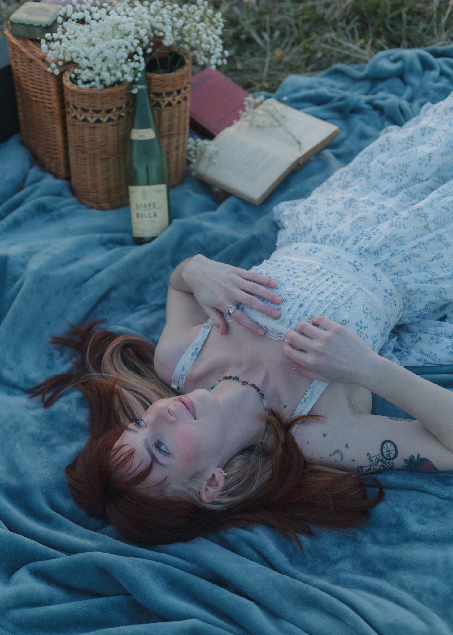 A red-haired woman in a floral dress lies on a blue blanket at a picnic with wine, books, and flowers.