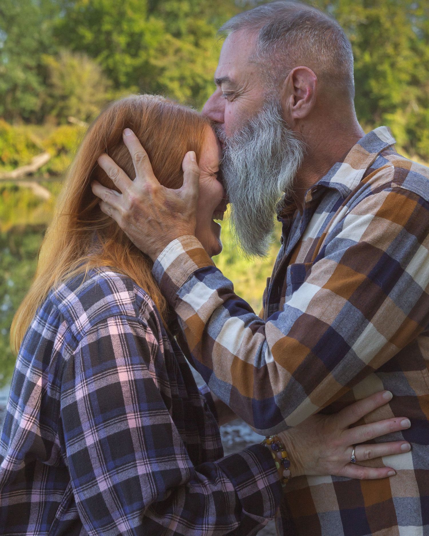 Two people in plaid shirts embrace in a romantic outdoor setting with sunlit trees in the background.Montgomery Count PA Couple Session