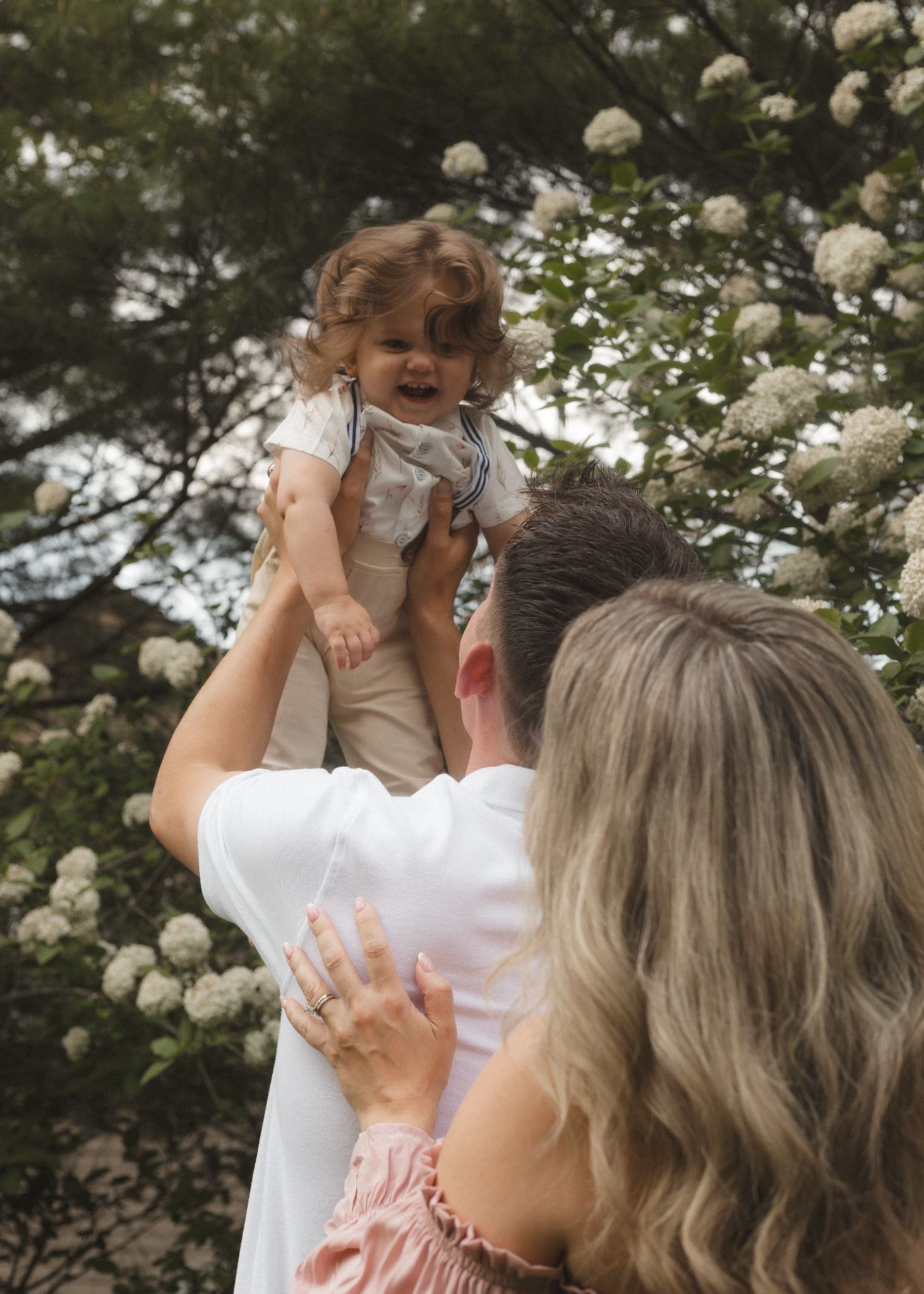 A woman holds a smiling baby up in the air outdoors near blooming white hydrangea bushes.