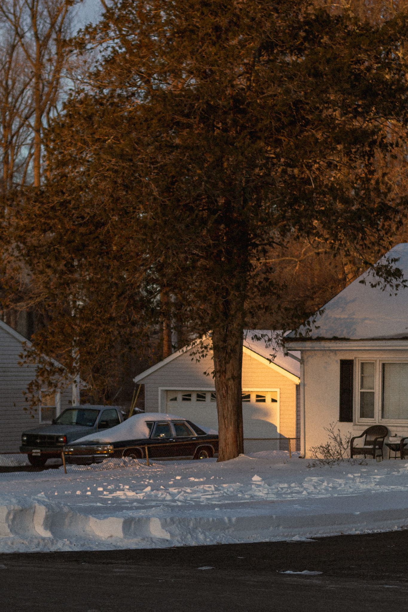 Snow covered suburban street at dusk with white houses and bare trees casting long shadows in winter.