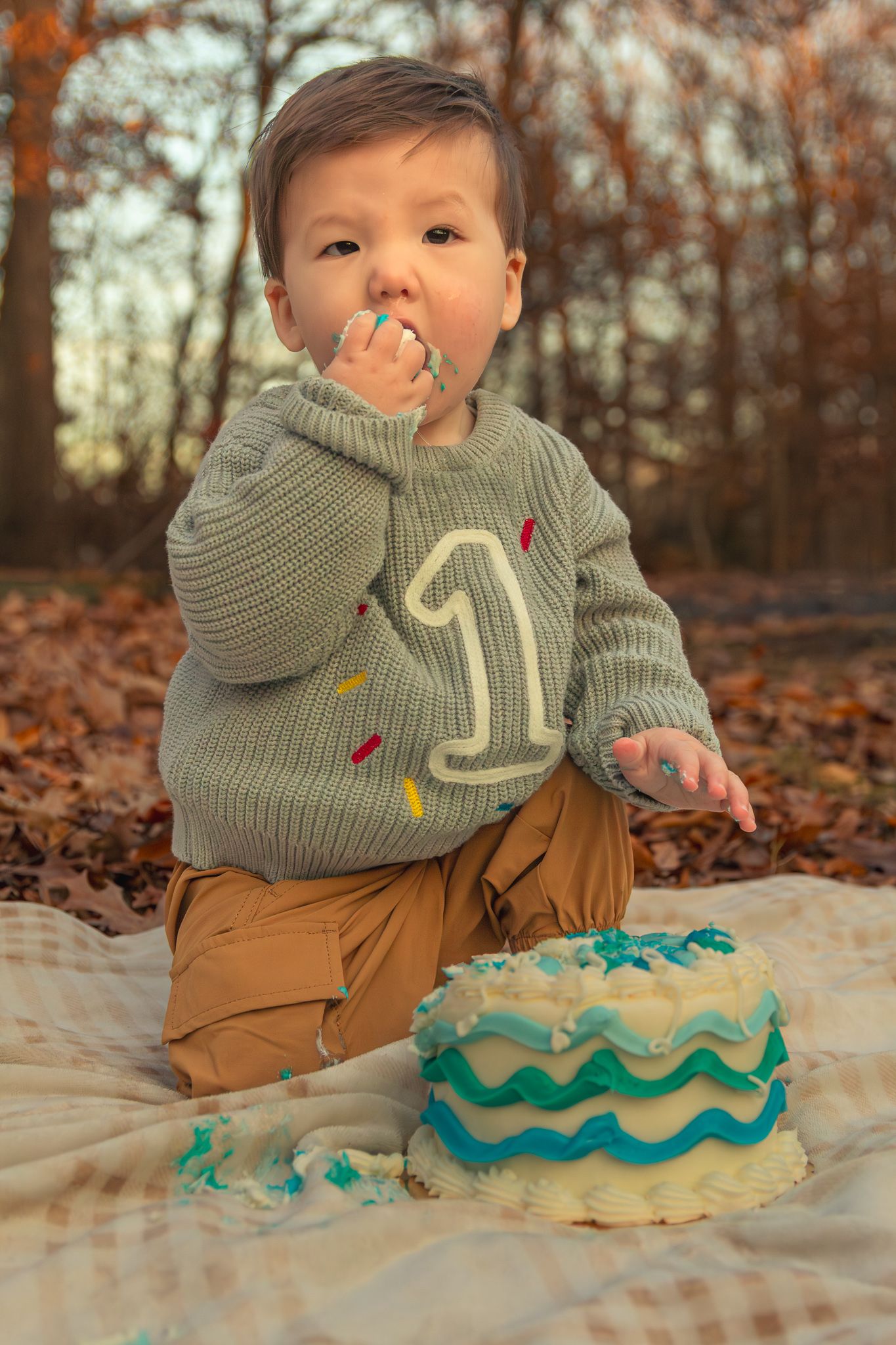 A small child in a gray sweater sits on a blanket with a turquoise birthday cake during autumn.