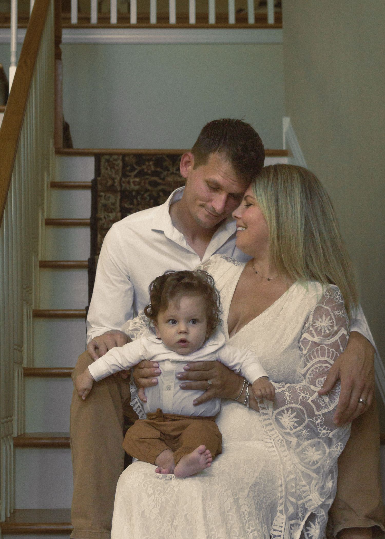 A couple shares tender moments with a baby while standing on wooden stairs in their home.