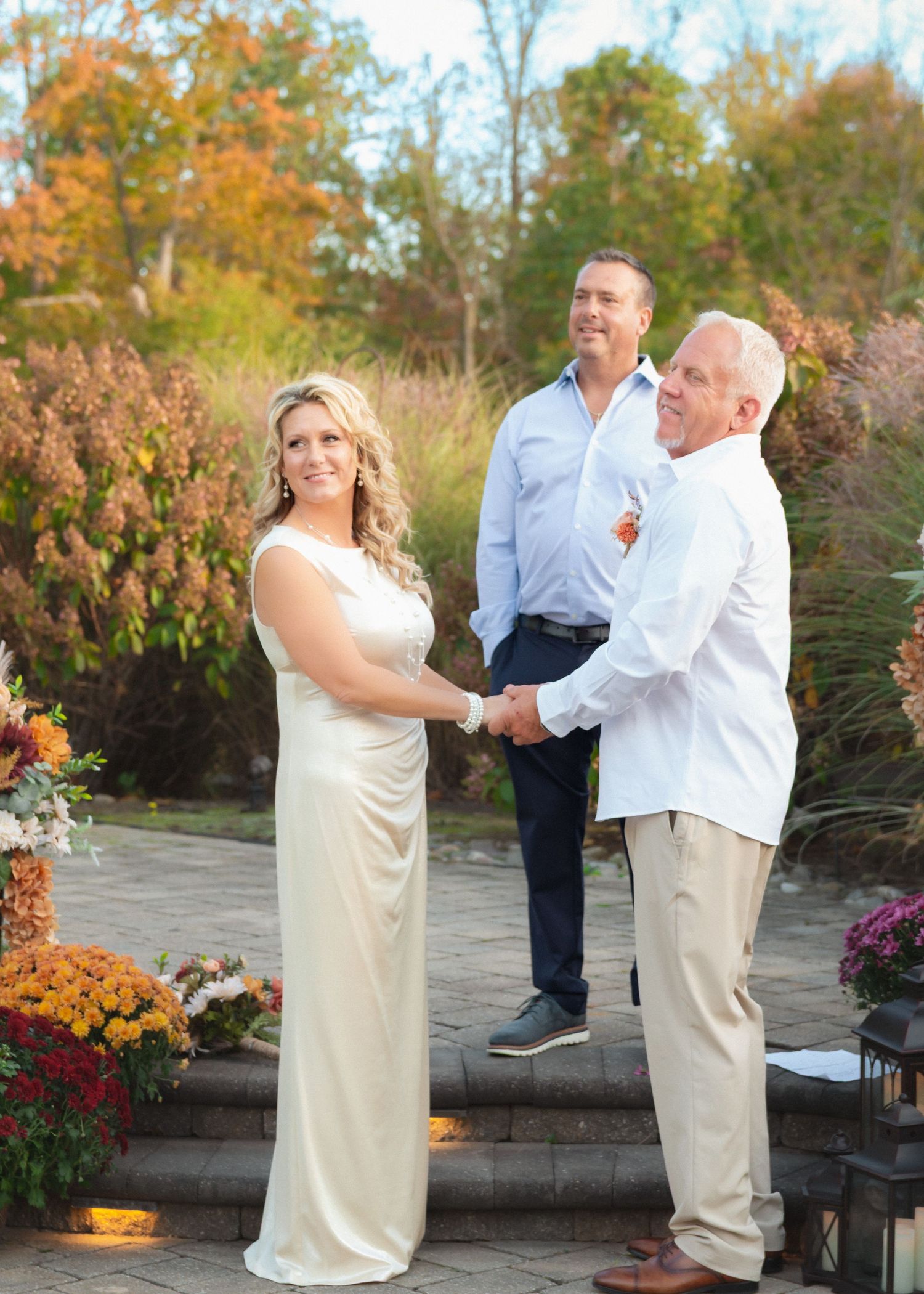 Outdoor autumn wedding ceremony on stone patio with colorful fall mums and ornamental grasses in background.