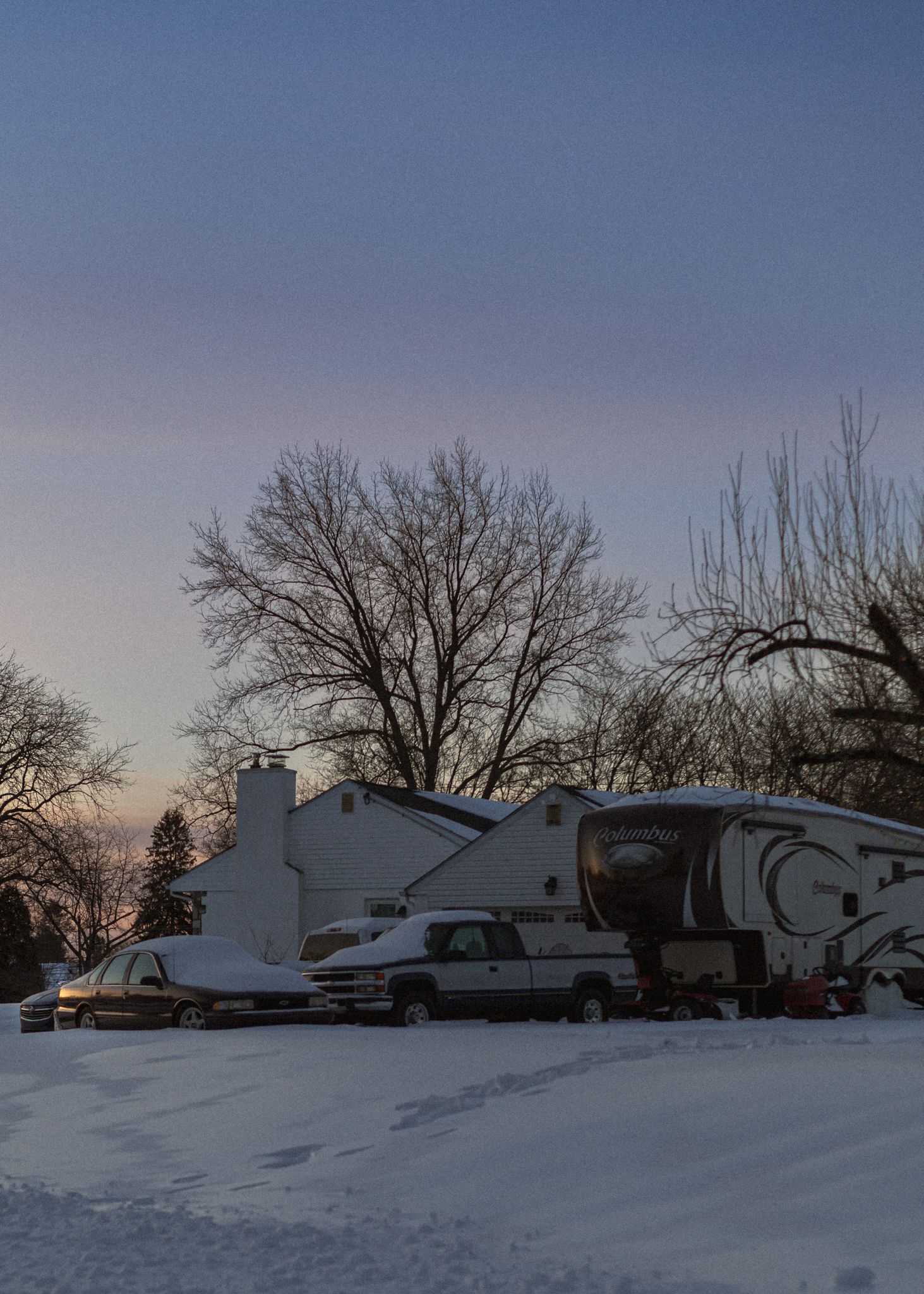 Snow covered RV campground with multiple trailers parked in a row at sunset with bare winter trees in the background.