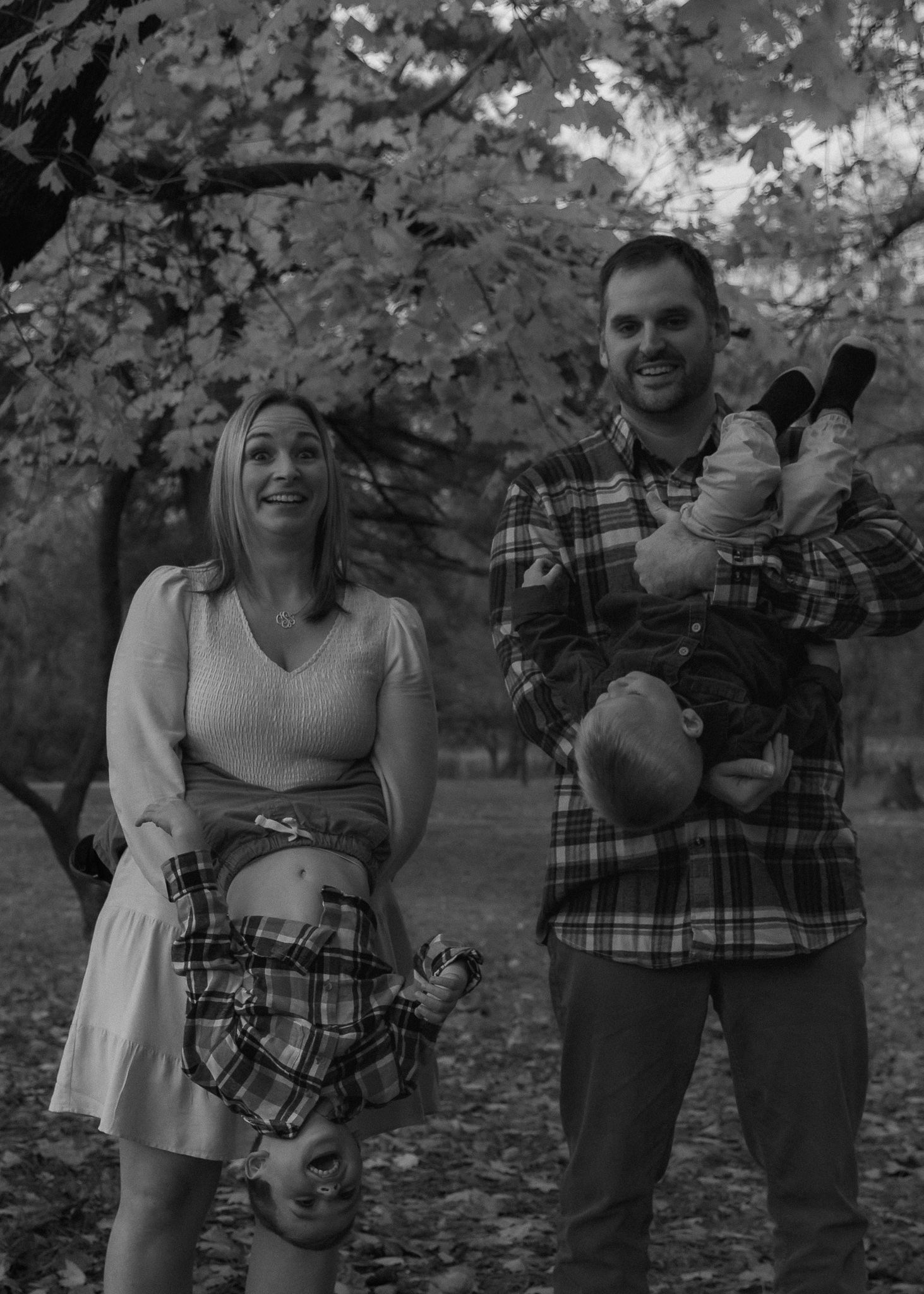 A couple playfully poses together in a park setting during autumn with fallen leaves around them in black and white.