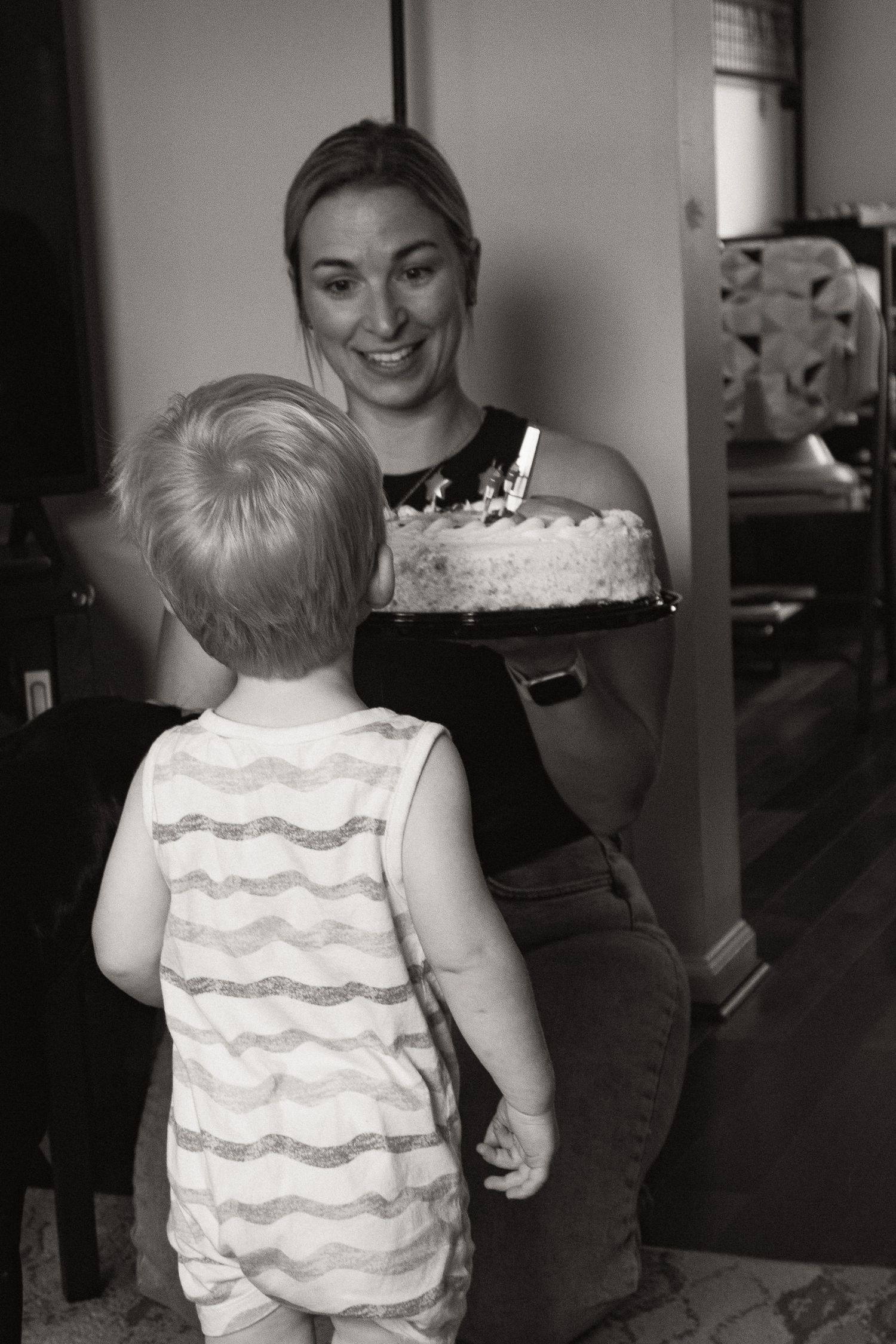 A toddler in a striped tank top watches someone holding a cake in this black and white photo series.