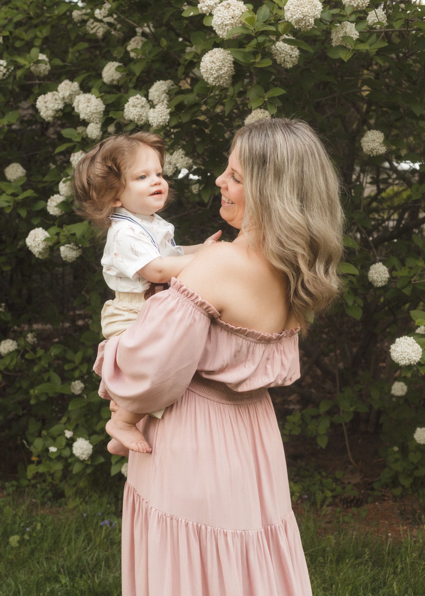A woman in a blush off-shoulder gown holds a toddler boy near white hydrangea bushes outdoors.