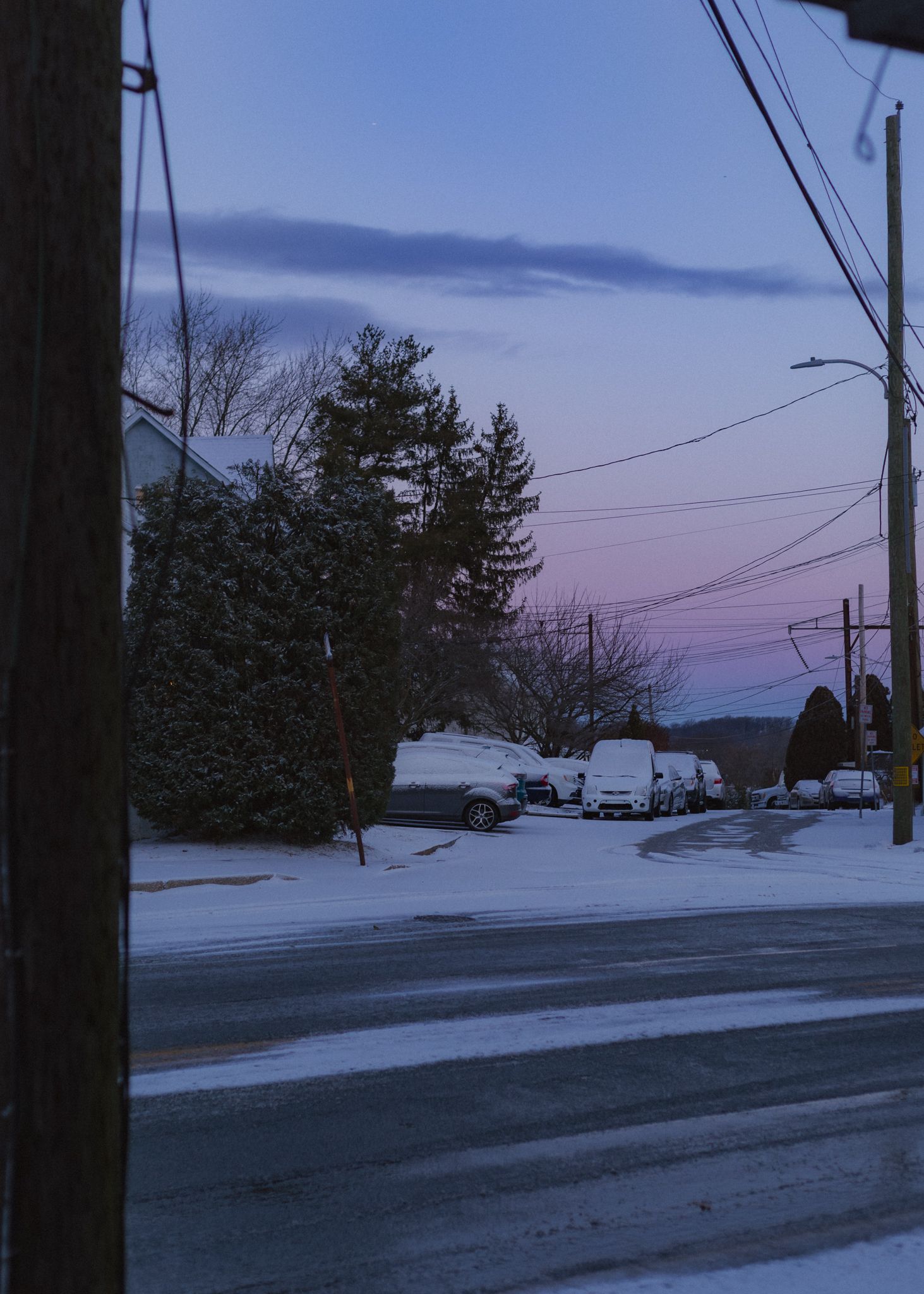 A snowy suburban street at dusk with power lines and evergreen trees silhouetted against a purple-blue winter sky.