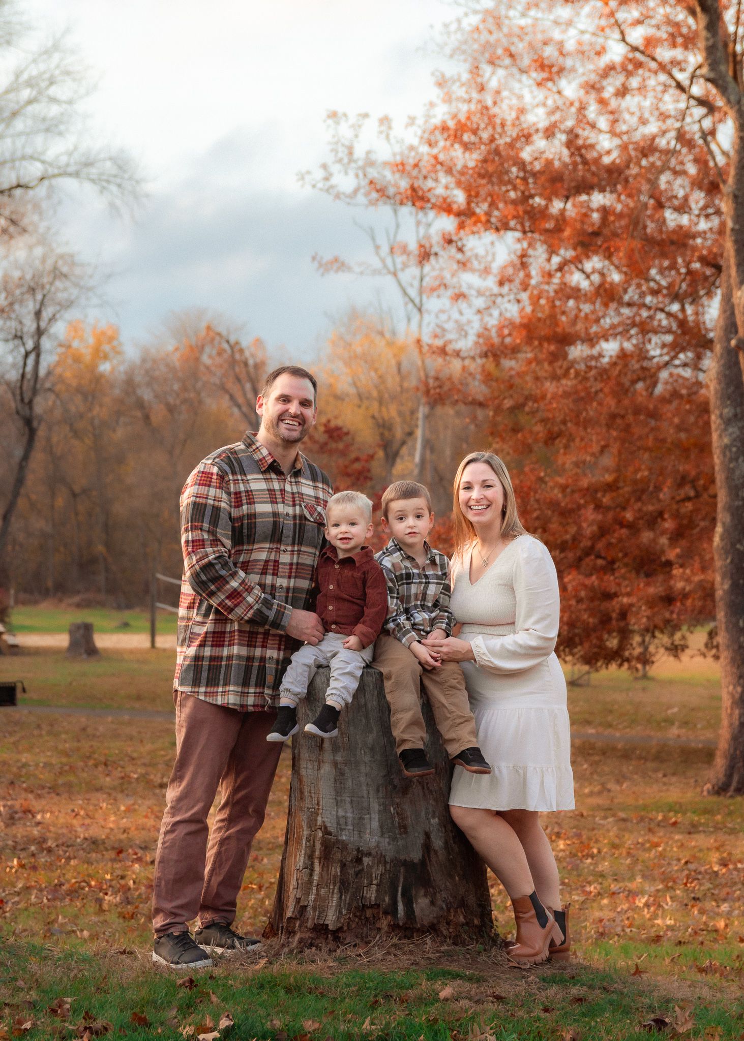 Family poses together in autumn setting with fallen orange leaves and tree stump in outdoor park setting.