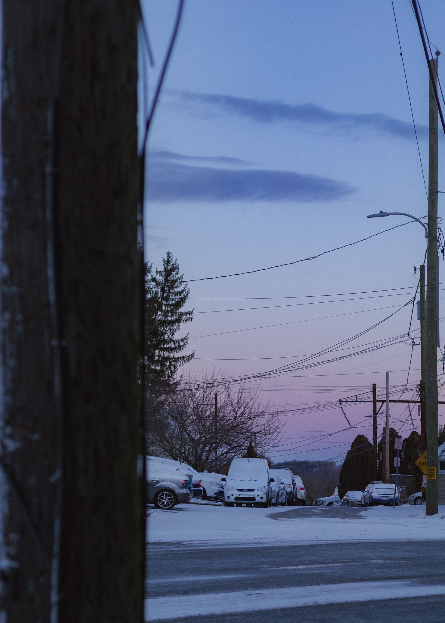 Snow covered residential street at dusk with power lines silhouetted against a purple and blue winter sky.