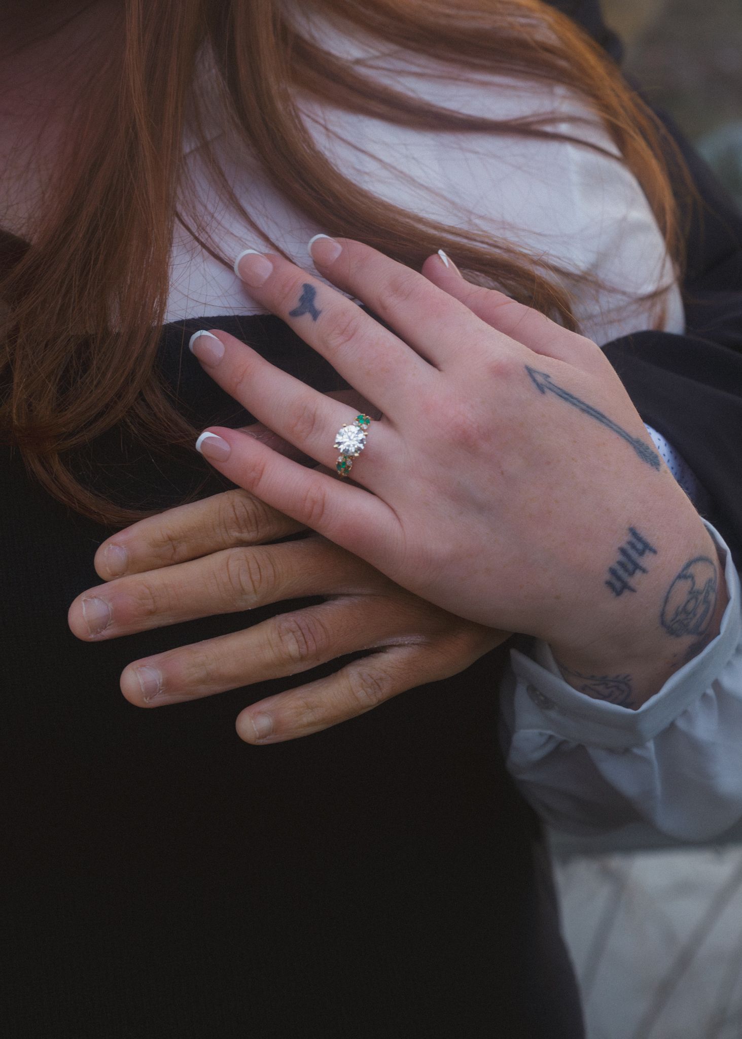 Close-up of a tattooed hand wearing a gold ring resting on a chest, with red hair visible in background.