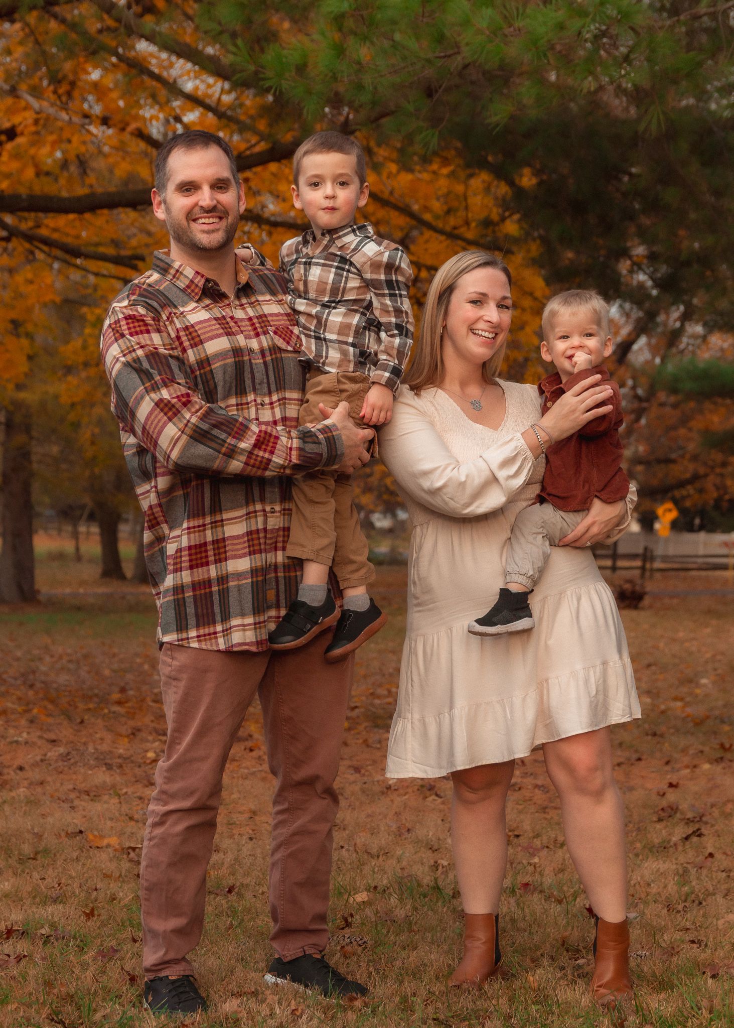 Family wearing coordinating plaid and cream outfits poses playfully in autumn setting with fallen leaves.