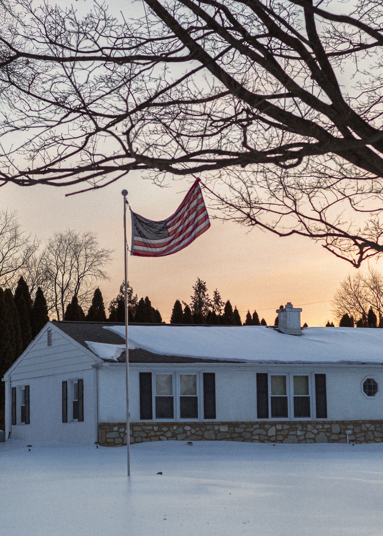 Panoramic winter view of a snow-covered ranch house with an American flag against a sunset sky framed by bare tree branches.