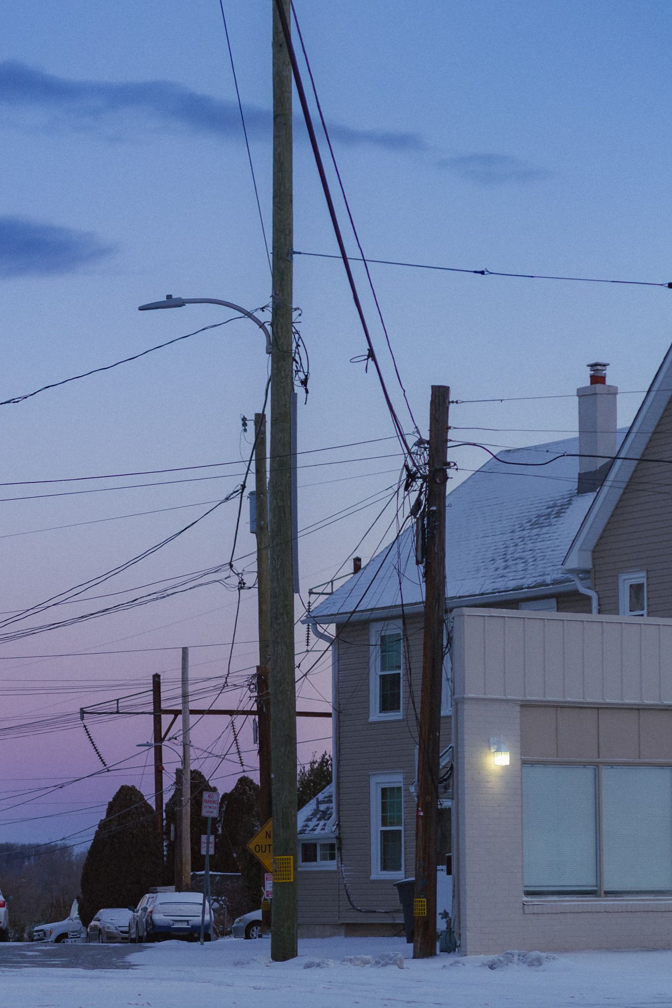 A snowy residential street at dusk with utility poles and power lines silhouetted against a purple-blue winter sky.