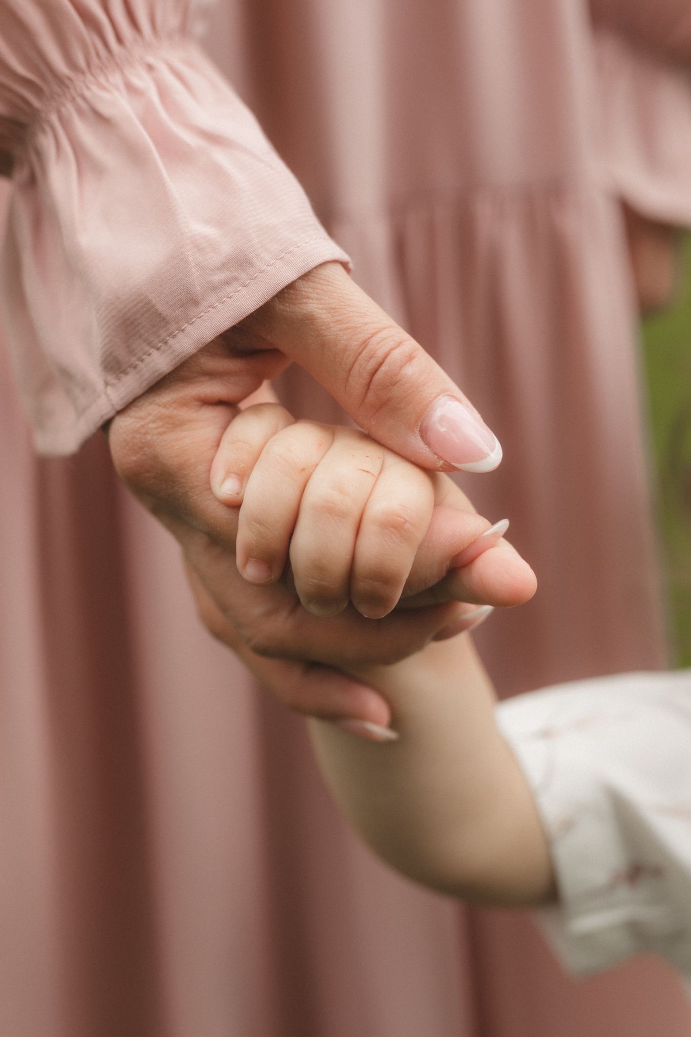 Close-up of a mother gently holding her young child's small hand, both wearing soft pink clothing outdoors.