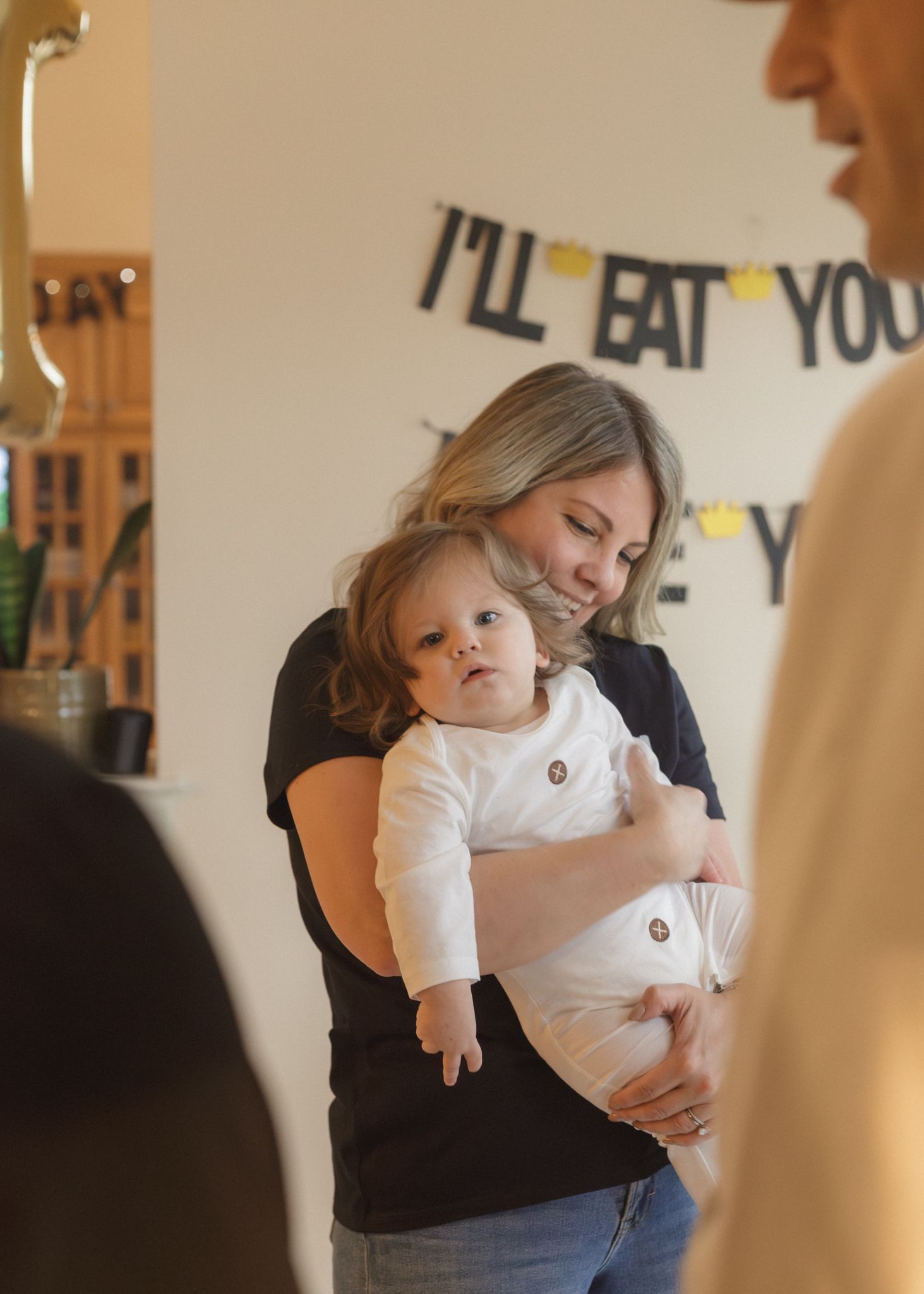 Woman holding baby at a birthday party with 'I'll Eat You Up' banner decoration in background.