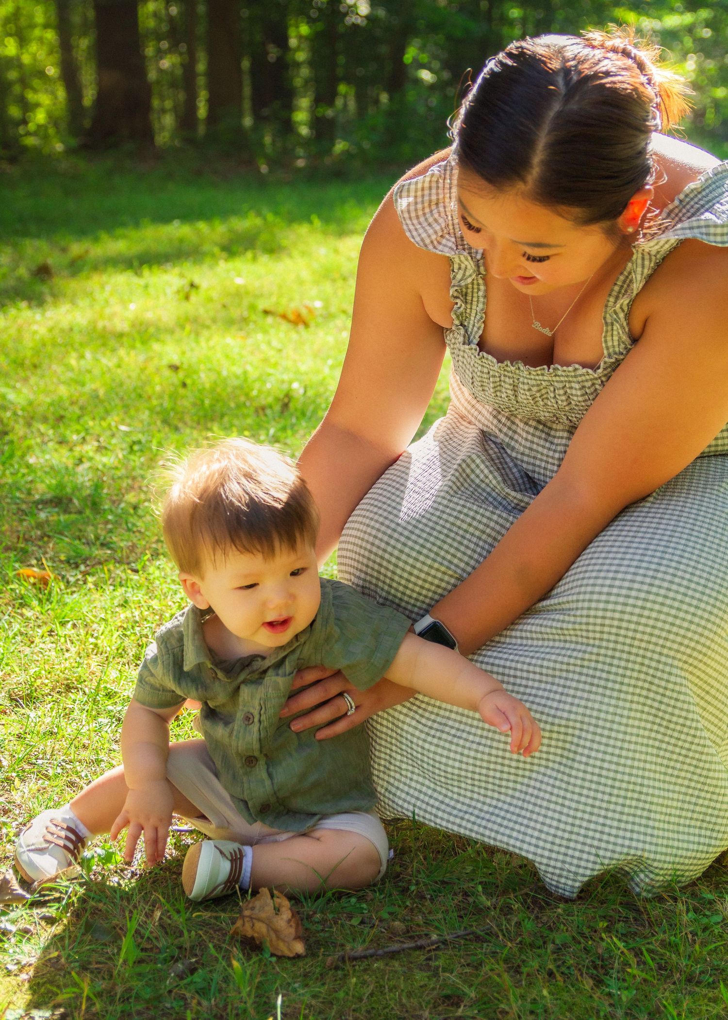 A tender moment captured on a sunny day as a caregiver in a gingham dress sits with a small child on green grass.