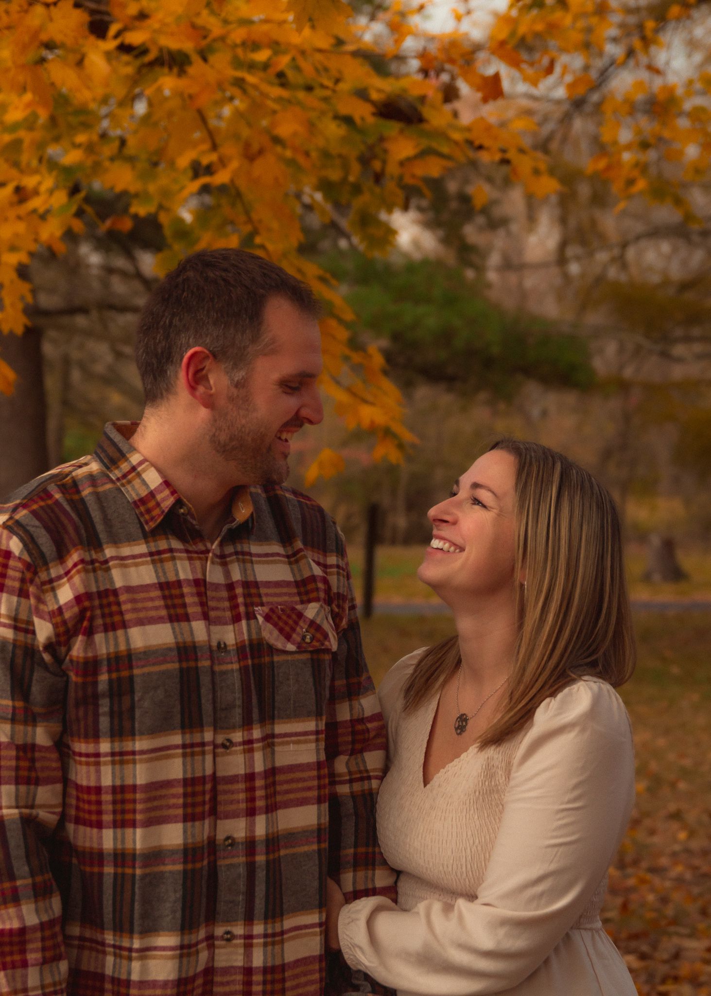 A couple shares joyful laughter while embracing in a park during autumn with orange leaves in the background.